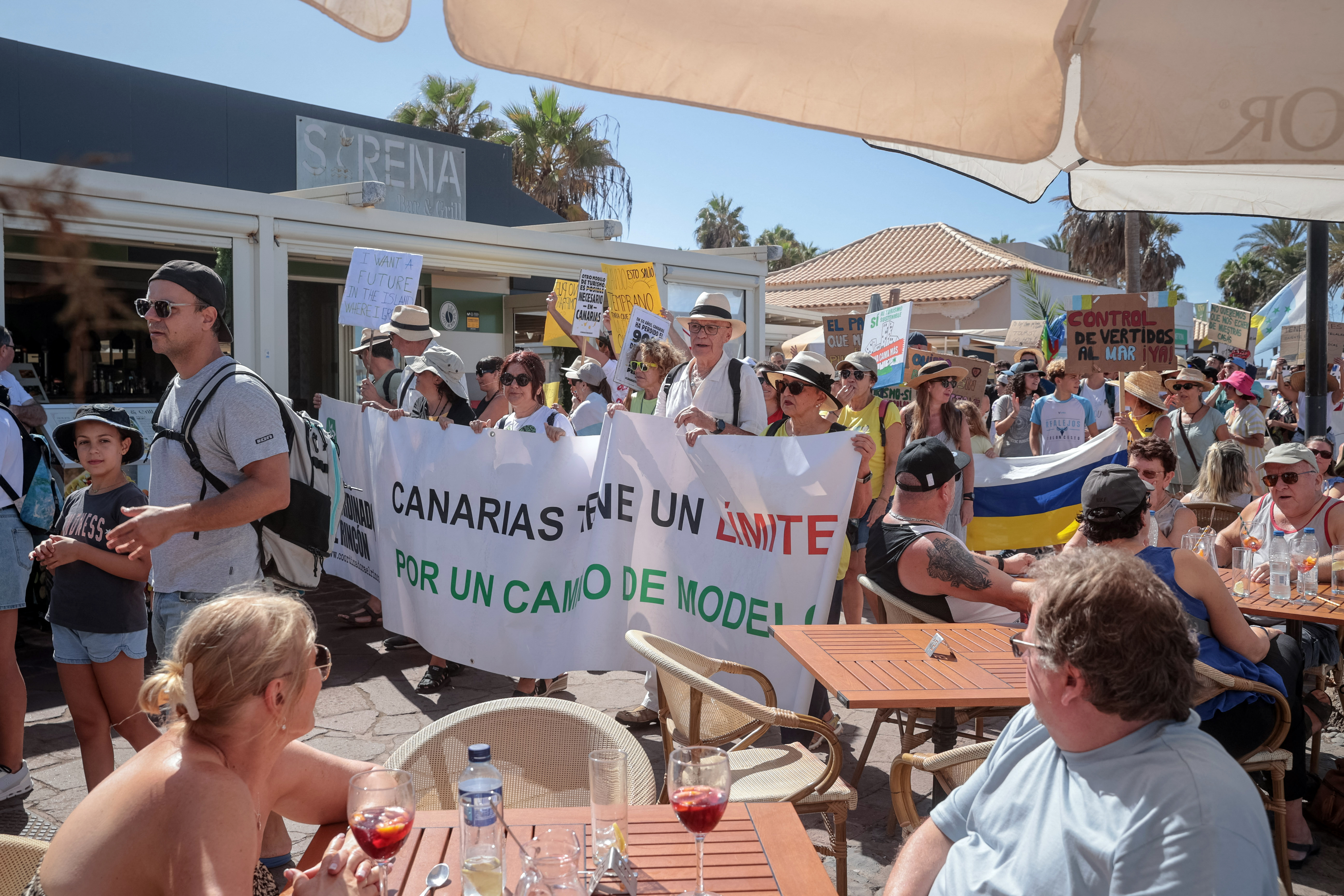 Tourists in a terrace look at protesters during a demonstration against mass tourism, in Arona on the Spanish Canary island of Tenerife, on October 20, 2024.