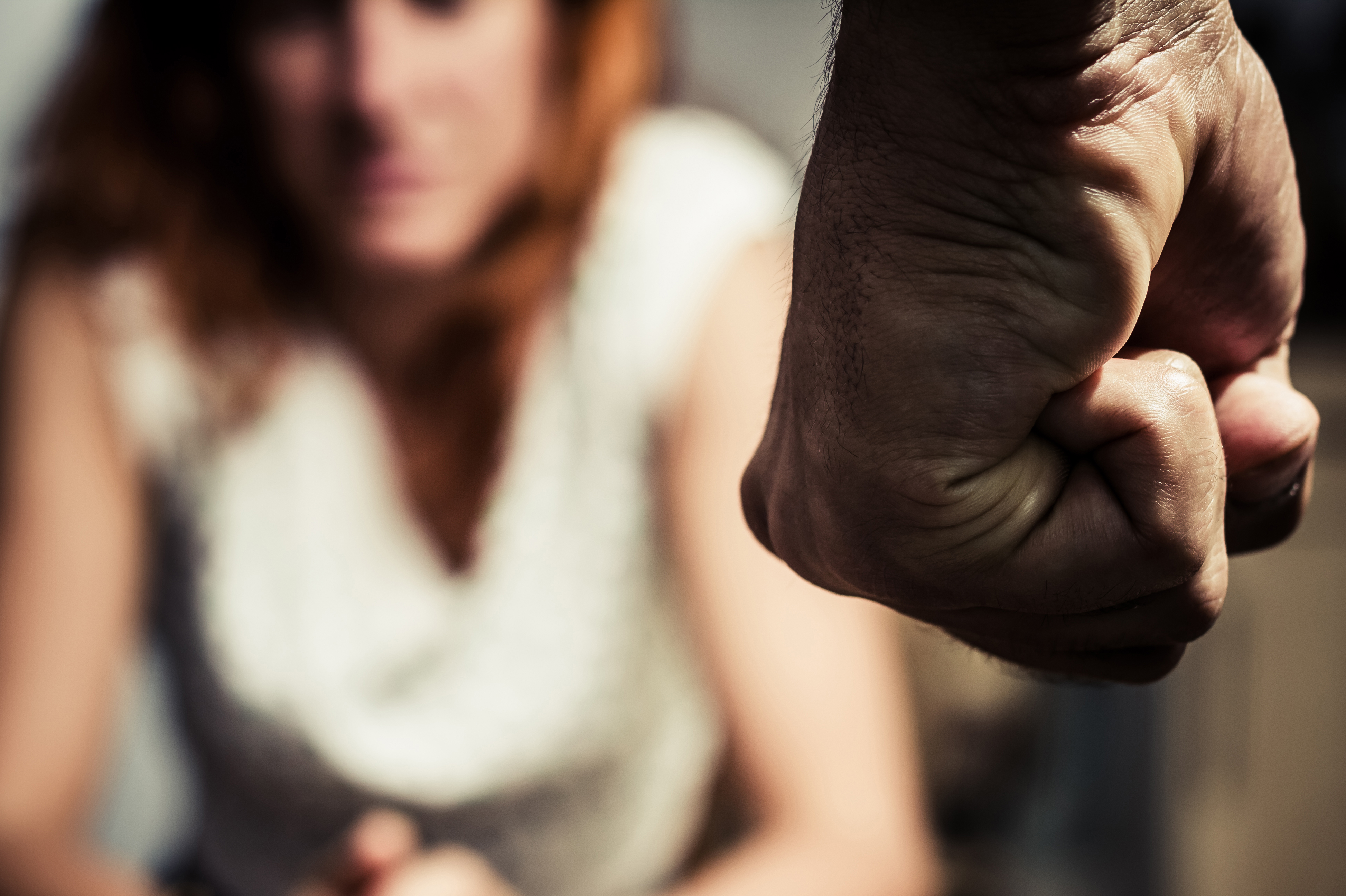 Young woman is sitting hunched at a table at home, the focus is on a man's fist in the foregound of the image