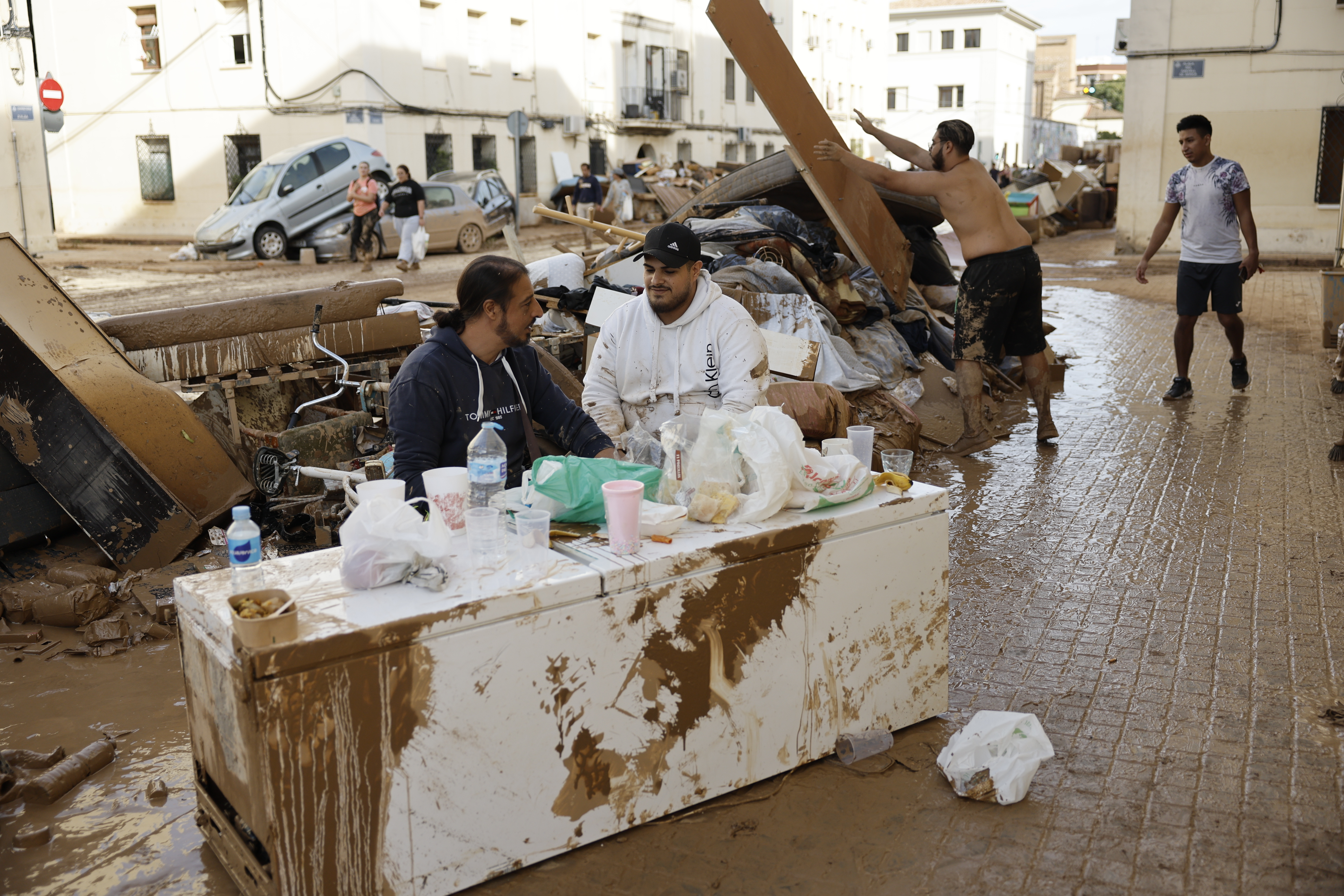 VALENCIA, 01/11/2024.- Vecinos del barrio de La Torre, comen en una mesa formada por una nevera en Valencia, este viernes. La búsqueda de desaparecidos, la identificación de víctimas mortales, las tareas de limpieza y la reparación de infraestructuras continúan tres días después de las inundaciones que han asolado la provincia de Valencia, en una jornada en la que el Gobierno envía a 500 militares más, que se sumarán a las 1.200 efectivos de la Unidad Militar de Emergencias (UME), para actuar en Utiel, Requena, Riba-roja, Torrent, Paiporta y Algemesí. EFE/ Biel Alino