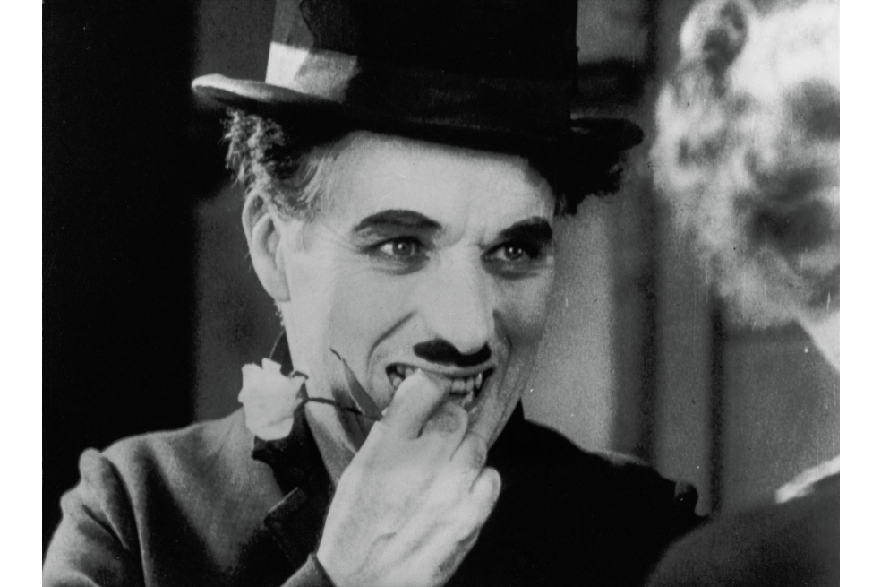 Primer plano de Charles Chaplin. La foto a blanco y negro muestra a Chaplin con un sombrero, su bigote insignia y los dedos de su mano derecha puestos sobre sus labios. Con ellos sostiene una flor.