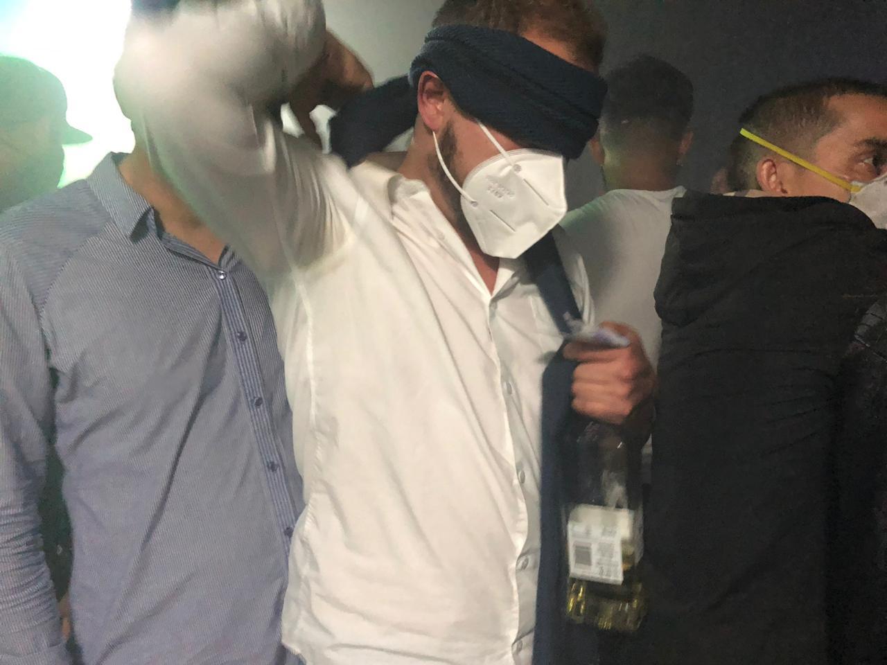 Un hombre con tapabocas y los ojos tapados sostiene una botella de Whisky.