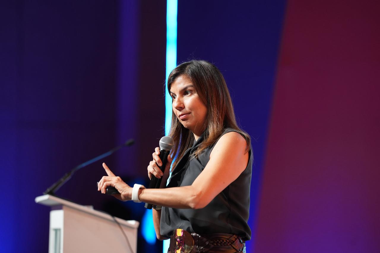 Ana Maiguashca, presidenta del Consejo Privado de Competitividad, de pie, sostiene un micrófono con la mano derecha y apunta con al izquierda hacia arriba mientras brinda un discurso.