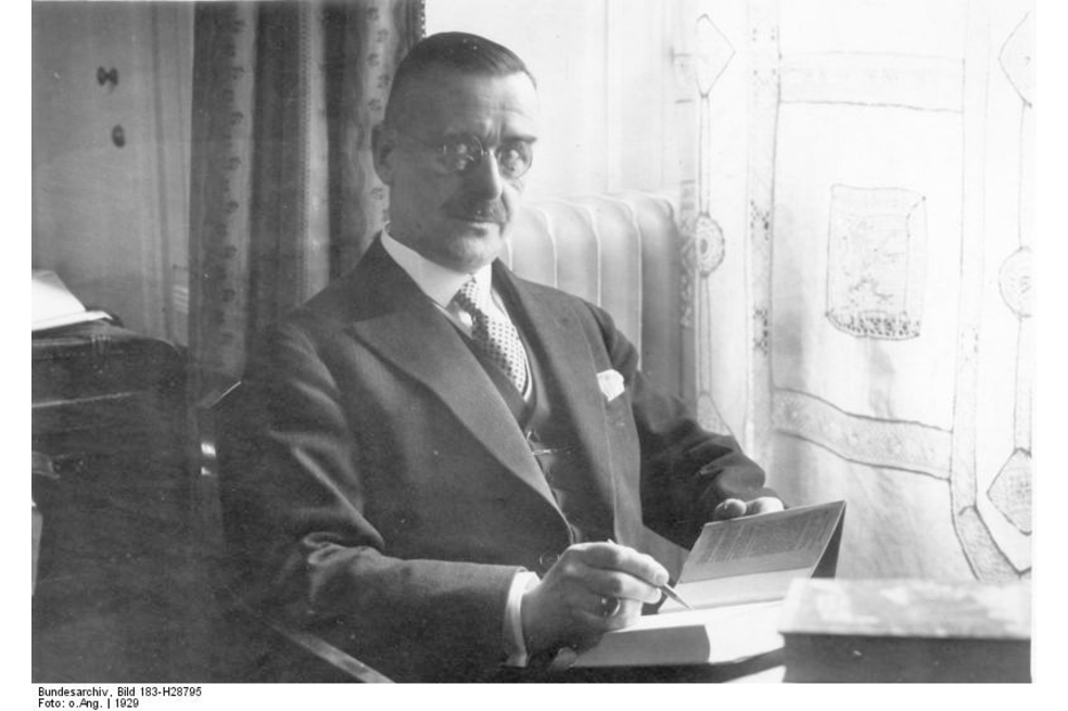 Thomas Mann aparece sentado en una habitación. En sus manos tiene un libro abierto y un esfero. La fotografía está a blanco y negro.