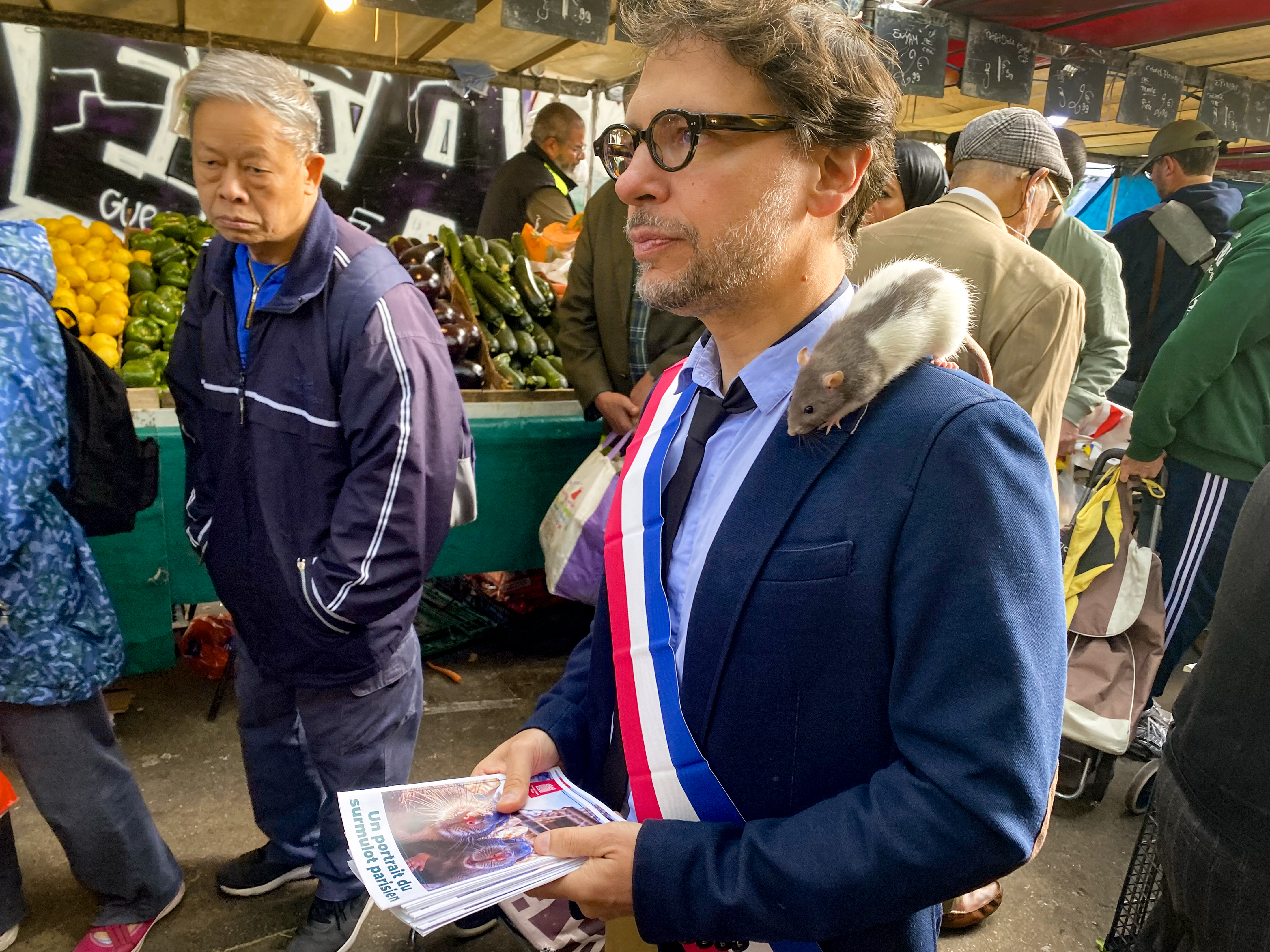 The rat 'Plume' sits on the shoulder of Gregory Moreau, the deputy mayor of Paris's 11th district, as he strolls through a market in the Belleville neighborhood in Paris on September 16, 2025. Deputy mayor Moreau has made it his mission to reconcile Parisians with their unloved co-inhabitants. (Photo by Ulrike KOLTERMANN / AFP)