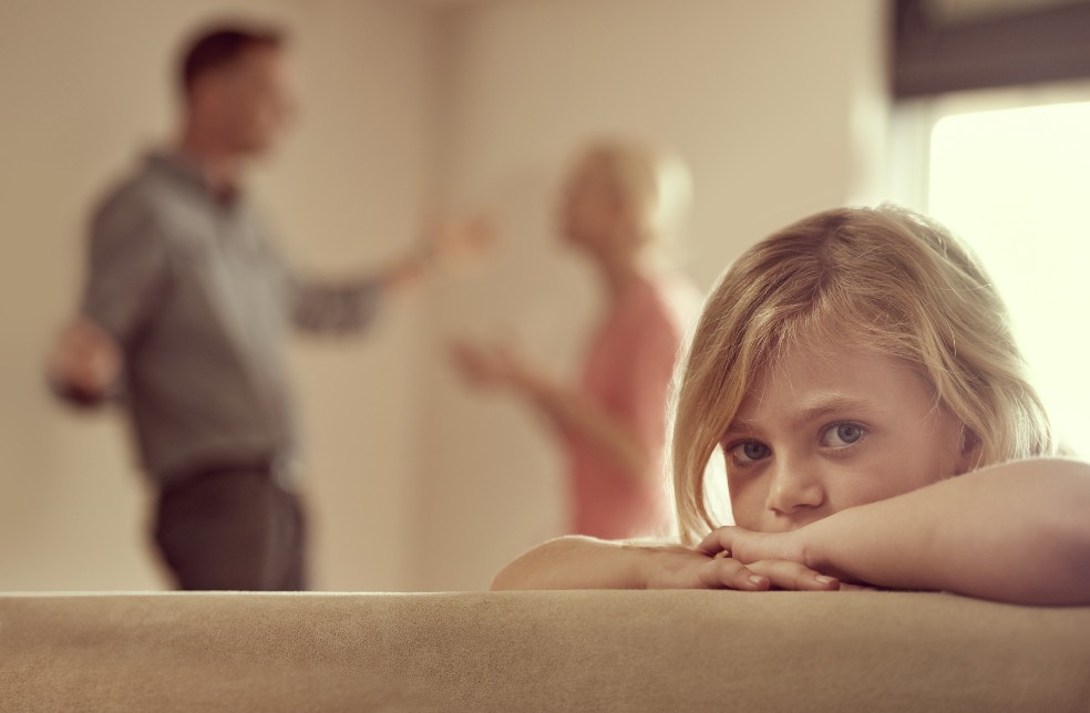 Dos padres discuten mientras su pequeña hija rubia les da la espalda sentada en un sofá.