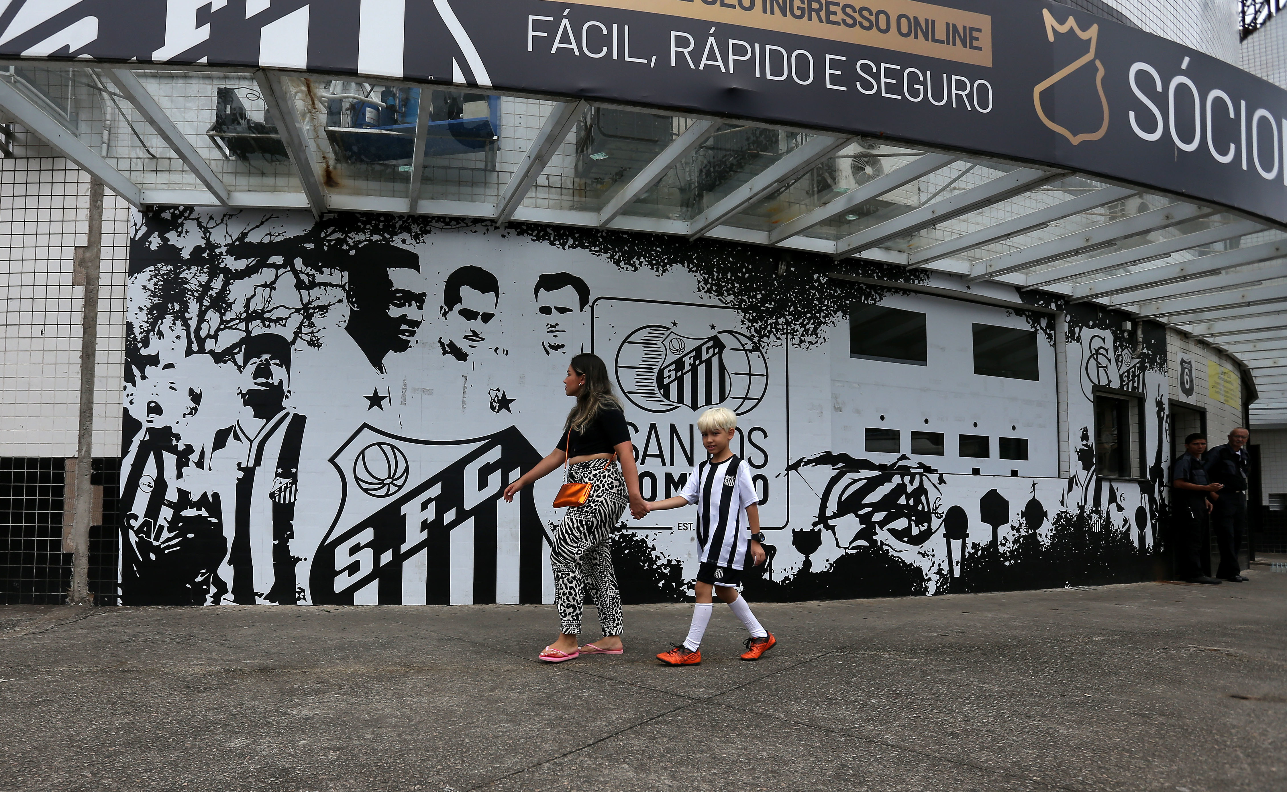 AME3199. SANTOS (BRASIL), 29/12/2022.- Una mujer y un niño caminan frente a un mural del Club Santos en el estadio Urbano Caldeira en la Vila Belmiro, tras conocerse la noticia de la muerte de la leyenda del fútbol brasileño Pelé, hoy, en la ciudad de Santos (Brasil). Los clubes brasileños de primera división sin excepción rindieron este jueves homenajes al tricampeón mundial Edson Arantes do Nascimento 'Pelé', que murió a sus 82 años y al que se refirieron como el "eterno" rey del fútbol. Como "Eterno" lo despidió el Santos, el club en que Pelé se inmortalizó y en que jugó casi toda su carrera. EFE/ Guilherme Dionizio