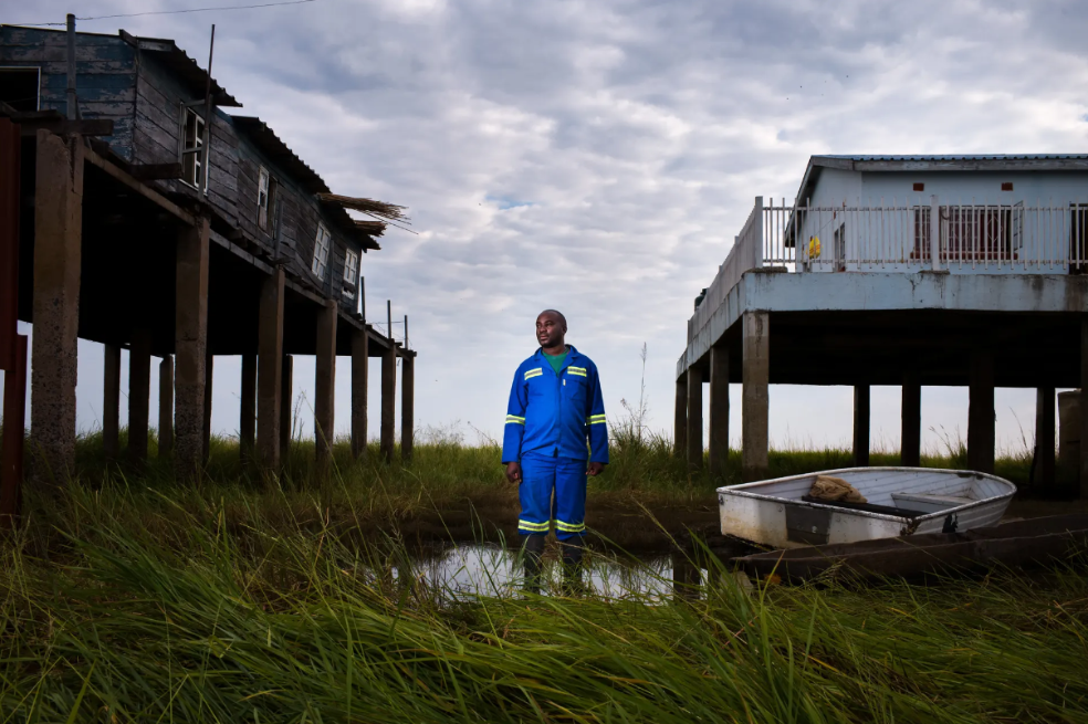 Las inundaciones perdidas: Un retrato de Lubinda Lubinda, jefe de estación de la Autoridad del Río Zambeze en Zambia. El bajo nivel de las aguas hace que su nueva casa (a la derecha) no tenga que ser construida tan alta. El pueblo barotse de la llanura aluvial está sometido a sequías más frecuentes debido al cambio climático y la deforestación.