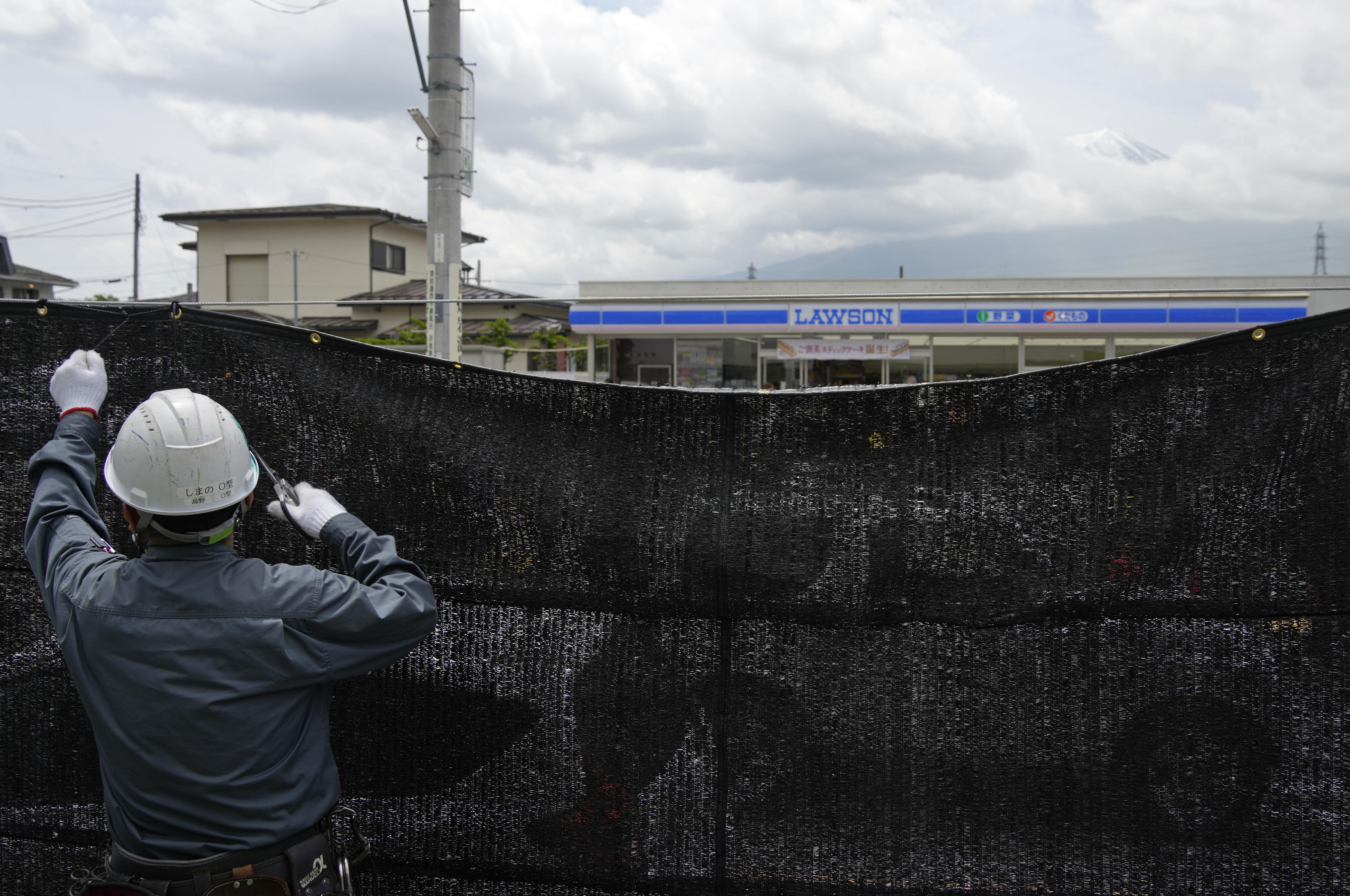 Fujikawaguchiko (Japan), 21/05/2024.- A worker installs a black shading net on the opposite side of the Lawson Kawaguchiko Ekimae convenience store as the summit of Mount Fuji is seen through the clouds in Fujikawaguchiko, north of Mount Fuji, Japan, 21 May 2024. The town of Fujikawaguchiko, north of Mount Fuji, installed a black shading net to block the view of Japan's iconic volcano from a particular spot before the Lawson Kawaguchiko Ekimae convenience store that became popular in the last two years to deter tourists who crowd there to photograph it following complaints from locals. (Japón) EFE/EPA/FRANCK ROBICHON