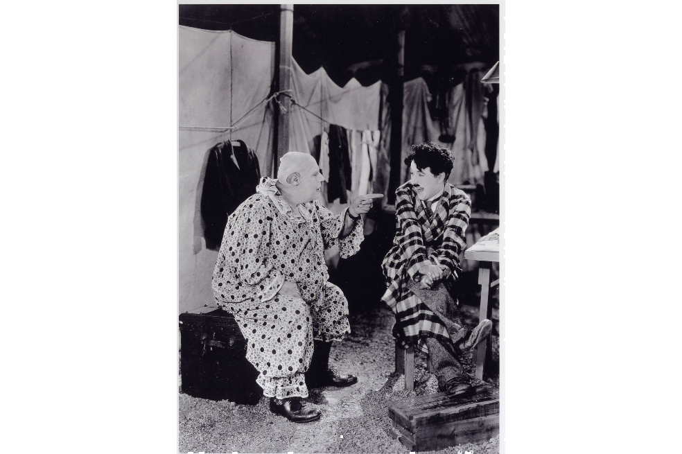 Charles Chaplin está sentado hablando con un personaje del circo. Chaplin tiene puesto un traje de cuadros.