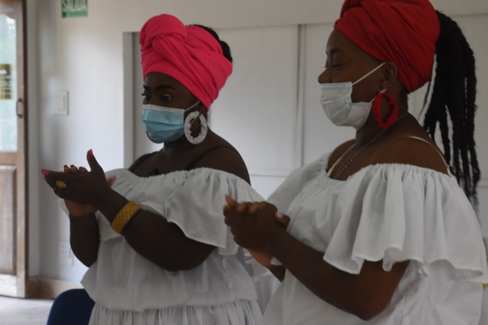 dos mujeres afros, vestidas con trajes típicos blancos y con tapabocas, cantan y aplauden.