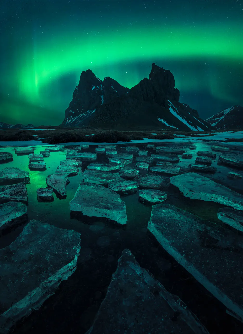 La imagen muestra la aurora boreal bailando, reflejada en un pequeño lago helado sobre la montaña Eystrahorn.