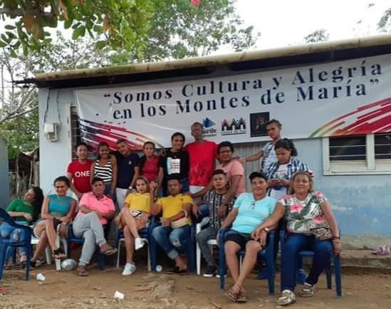 Foto de un grupo de hombres y mujeres de la población LGBT, sentados y de pie. Están en un entorno rural y detrás de ellos hay una pancarta que dice: "Somos cultura y alegría en los Montes de María".