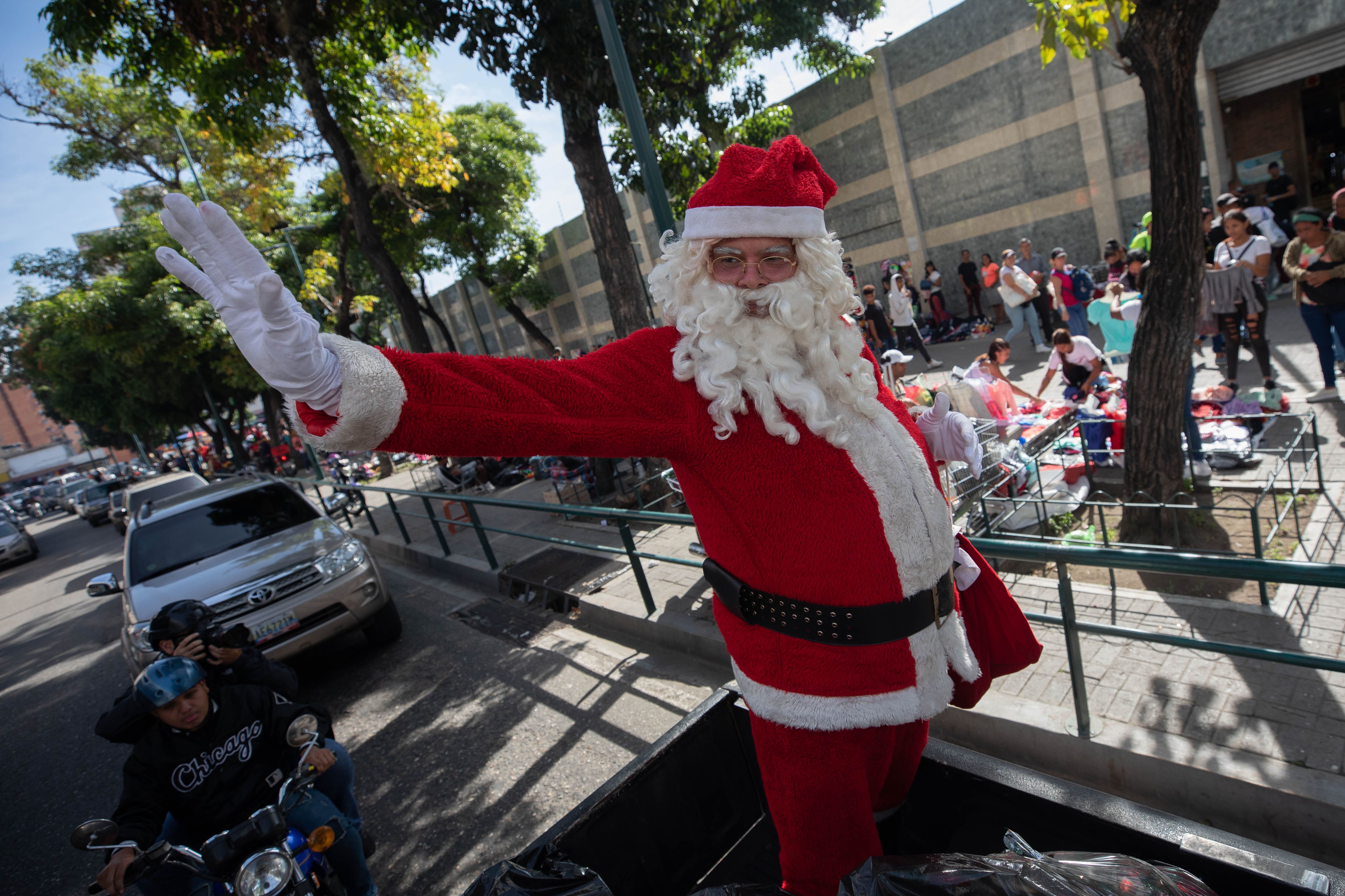 AME1260. CARACAS (VENEZUELA), 17/12/2022.- Un hombre vestido de Santa Claus saluda mientras recorre las calles desde un vehículo durante la entrega de regalos de la iniciativa "Un Juguete, una buena noticia", hoy, en Caracas (Venezuela). Un Santa Claus y varios voluntarios -la mayoría periodistas- entregaron este sábado regalos a 350 niños de una comunidad de Caracas, gracias a donativos del gremio y de particulares, durante la iniciativa llamada "Un juguete, una buena noticia", que un grupo de trabajadores de la prensa desarrolla desde 2017 con el objetivo de arrancar sonrisas a los pequeños. EFE/ Rayner Peña R.