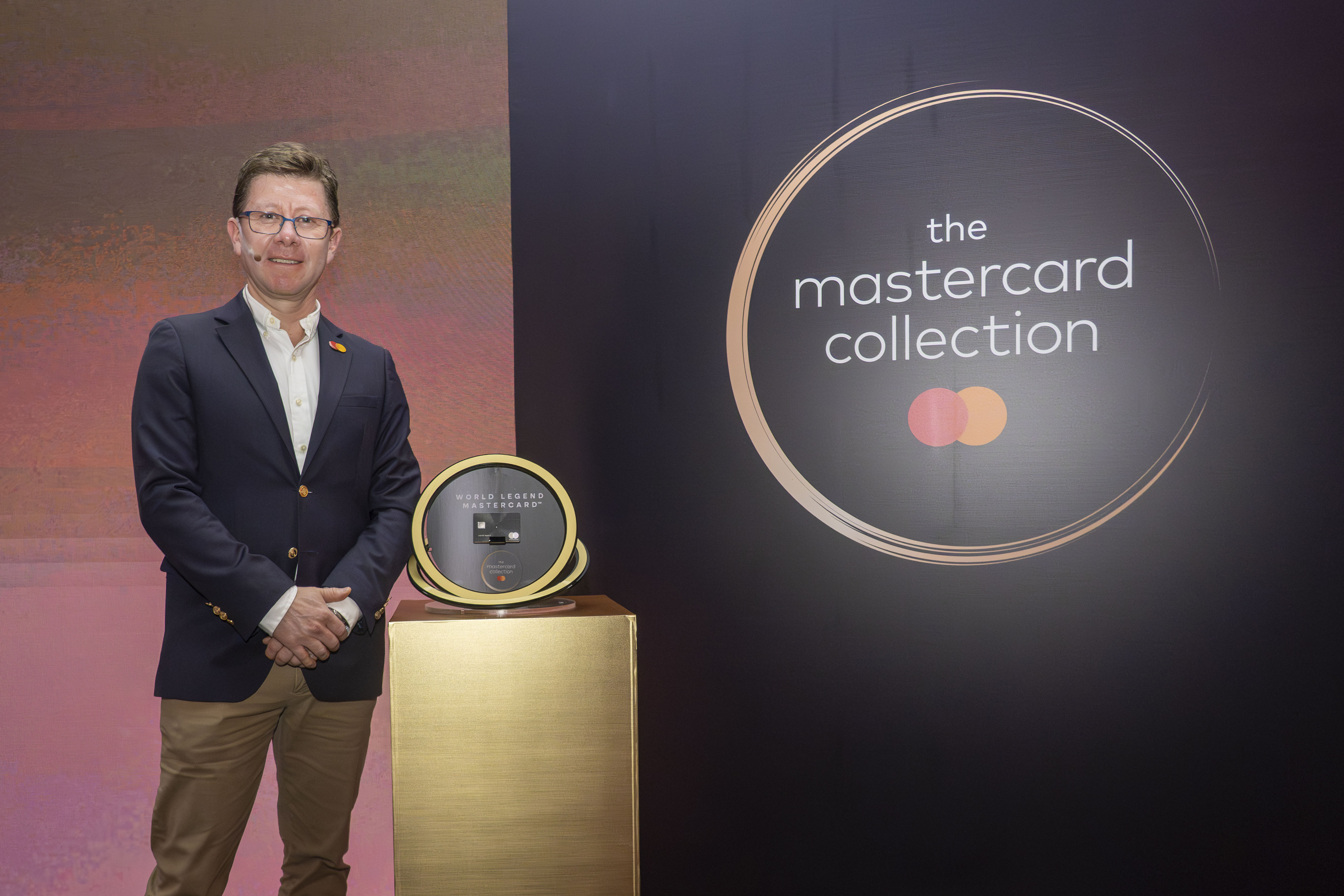 Fotografía de un evento de Mastercard con un display dorado que exhibe la tarjeta “World Legend Mastercard”. Un hombre con blazer azul y pantalón beige posa junto al pedestal, frente a un fondo con el logo de Mastercard y el texto “the mastercard collection”.