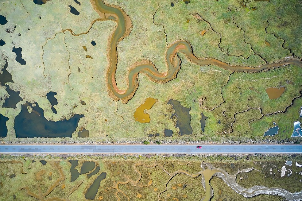 Categoría humedales: Javier Lafuente, de España, tomó esta foto con su dron. Lafuente se enfrentó a los desafíos de la luz solar reflejada por el agua y las cambiantes condiciones de luz. Dividiendo el humedal en dos, esta carretera se construyó en la década de 1980 para proporcionar acceso a una playa.