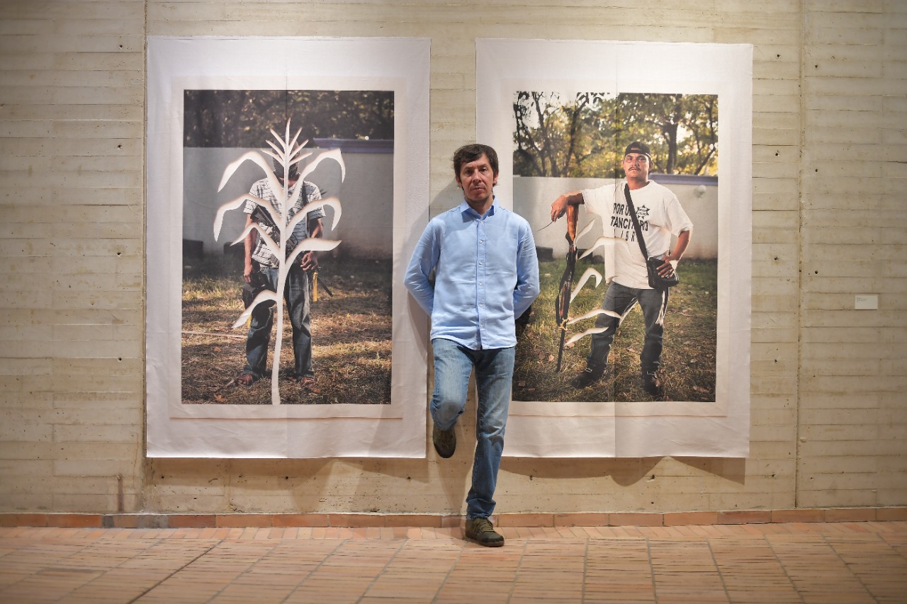 En el centro de la foto Futuro Moncada, autor de “Venceremos”, acompañado de una de sus piezas: fotografías, intervenidas con una silueta de hoja de maíz, de dos miembros de las Autodefensas de México, que surgieron inicialmente como una forma de resistencia de la comunidad indígena de Michoacán ante los grupos del narcotráfico.