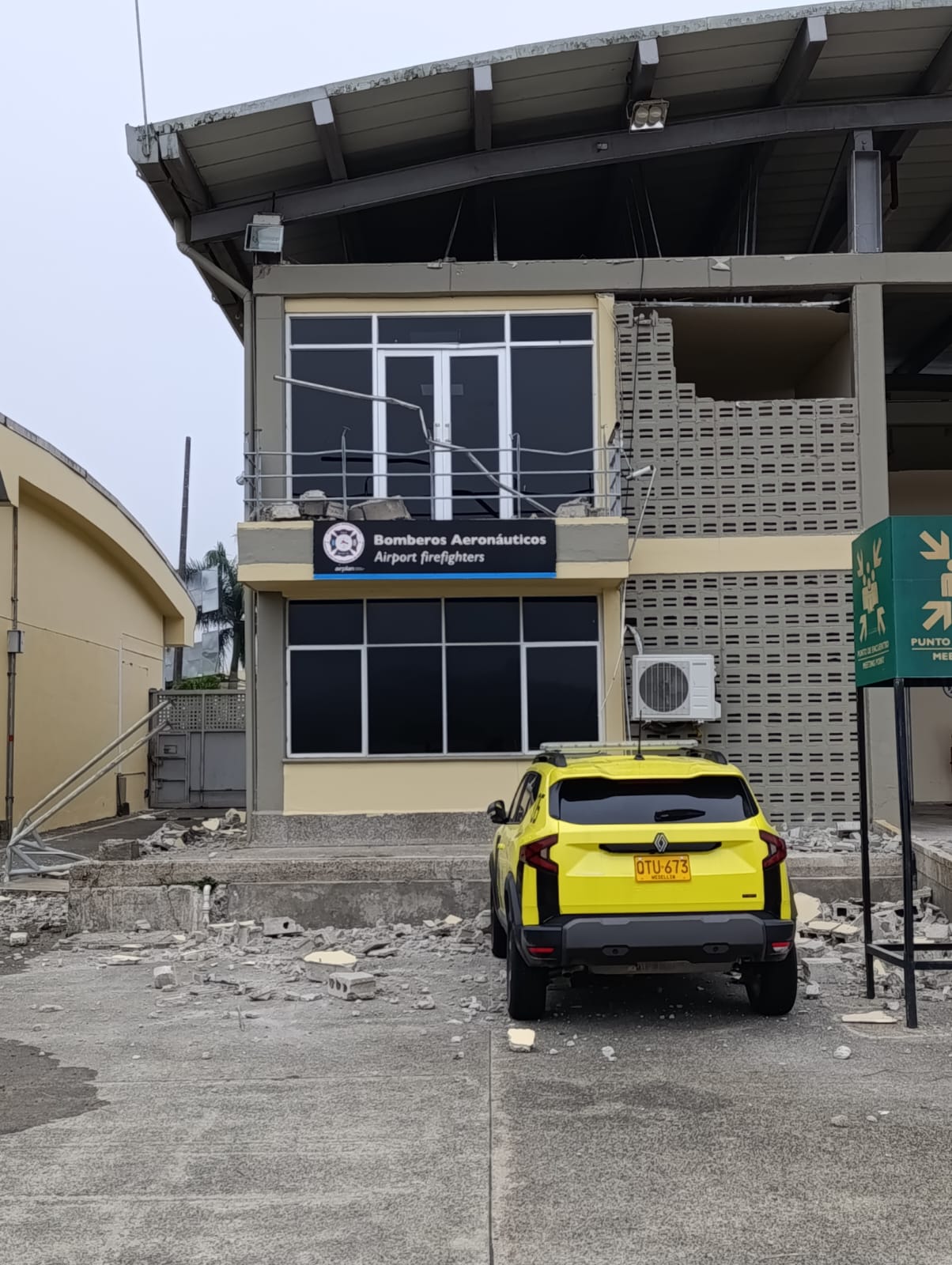 Edificio de dos pisos de los Bomberos Aeronáuticos con daños estructurales visibles: ventanas rotas en la parte superior, baranda metálica doblada y grietas en las paredes. En el suelo hay escombros y fragmentos de concreto frente a la entrada. Un automóvil amarillo con placa OTU-673 está estacionado junto a la fachada, rodeado de restos caídos. La escena refleja un inmueble de servicios de emergencia afectado por colapso parcial o evento sísmico.