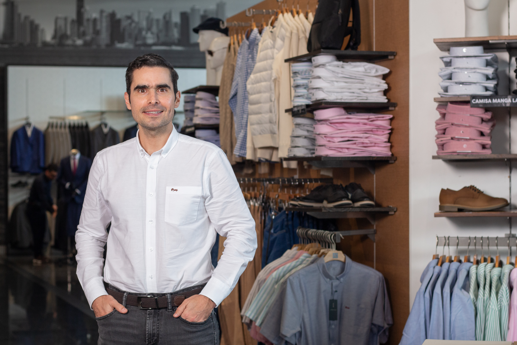 Esteban González de pie en tienda de Arturo Calle, con camisa blanca y pantalón oscuro, rodeado de estanterías con ropa ordenada.