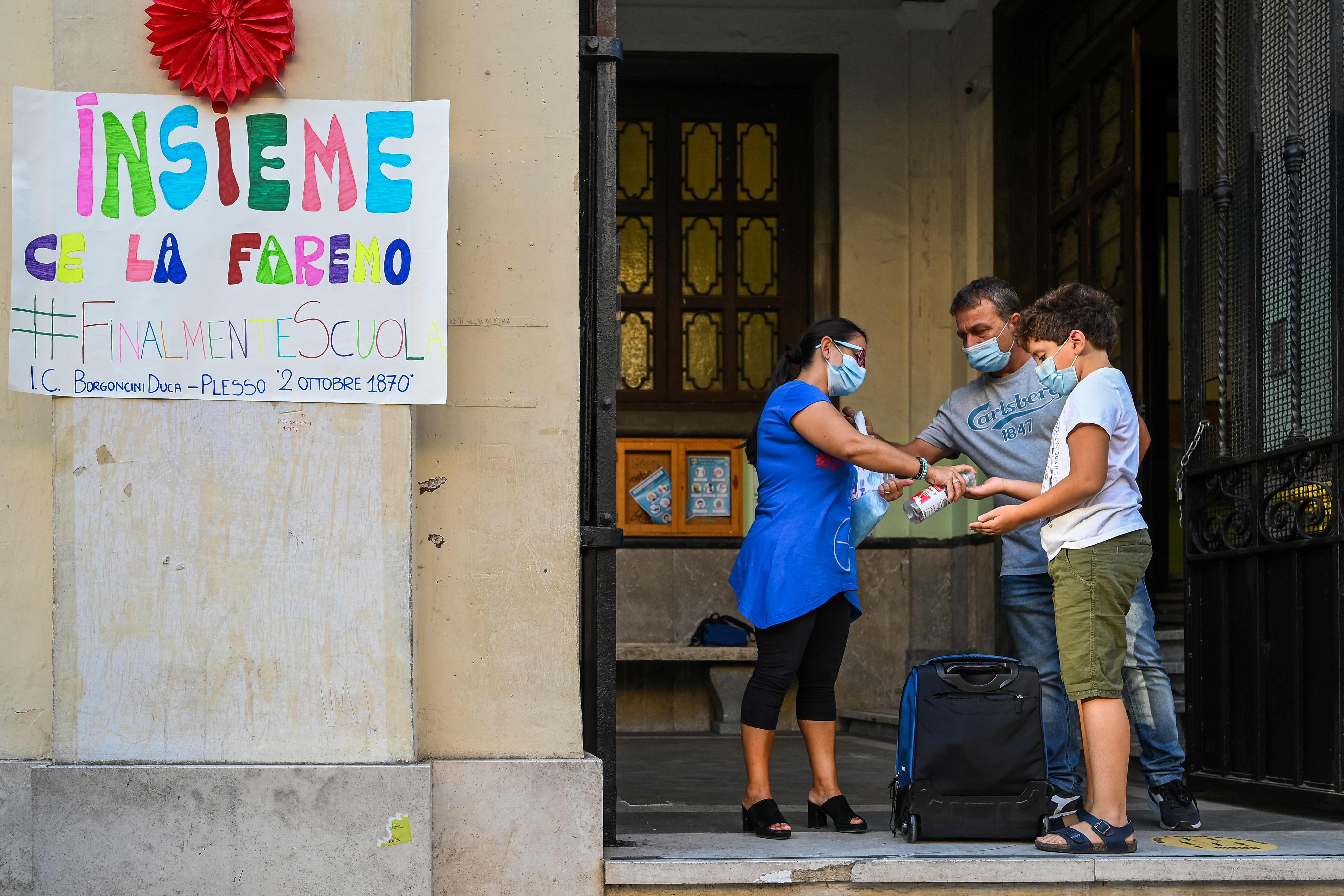 A shool staff (C) wearing a face mask dispenses hand sanitizer gel to a pupil (R), also wearing a face mask, arriving on September 14, 2020 for the start of the school year at the Luigi Einaudi technical high school in Rome, during the the COVID-19 infection, caused by the novel coronavirus. - Schools in some European nations were set to open on September 14, 2020 with millions returning to classrooms in Italy, Greece and Romania. The inscription (L) reads : "Together we will make it. #finally school'. (Photo by Vincenzo PINTO / AFP)