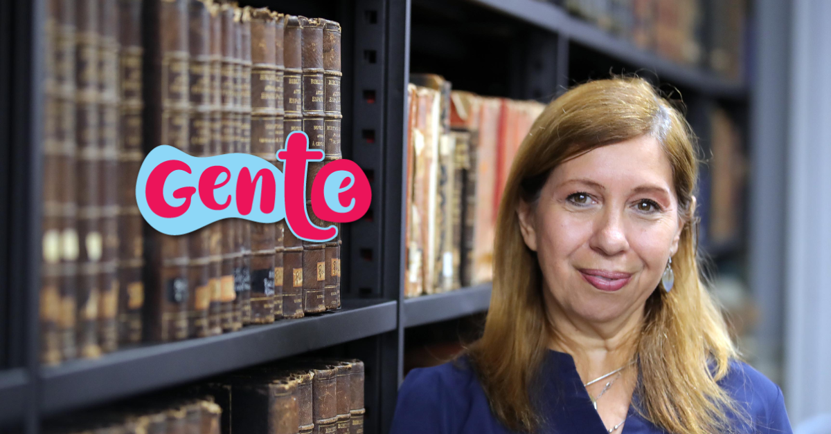 Retrato de una mujer de mediana edad, blanca, pelo rubio, blusa azul, reclinada al lado de una biblioteca llena de textos antiguos.