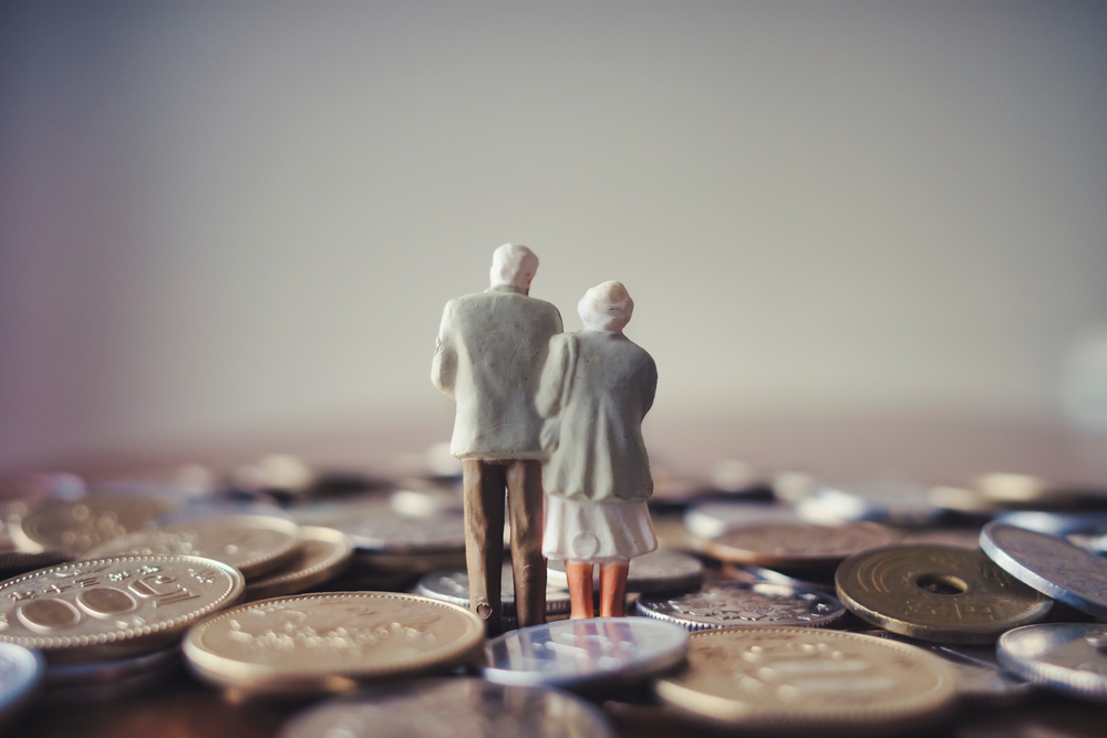 Figura en miniatura de una pareja de adultos mayores de espaldas, de pie sobre un fondo de monedas, representando el sistema de pensiones y la postergación de los fondos generacionales del ROP en Costa Rica.