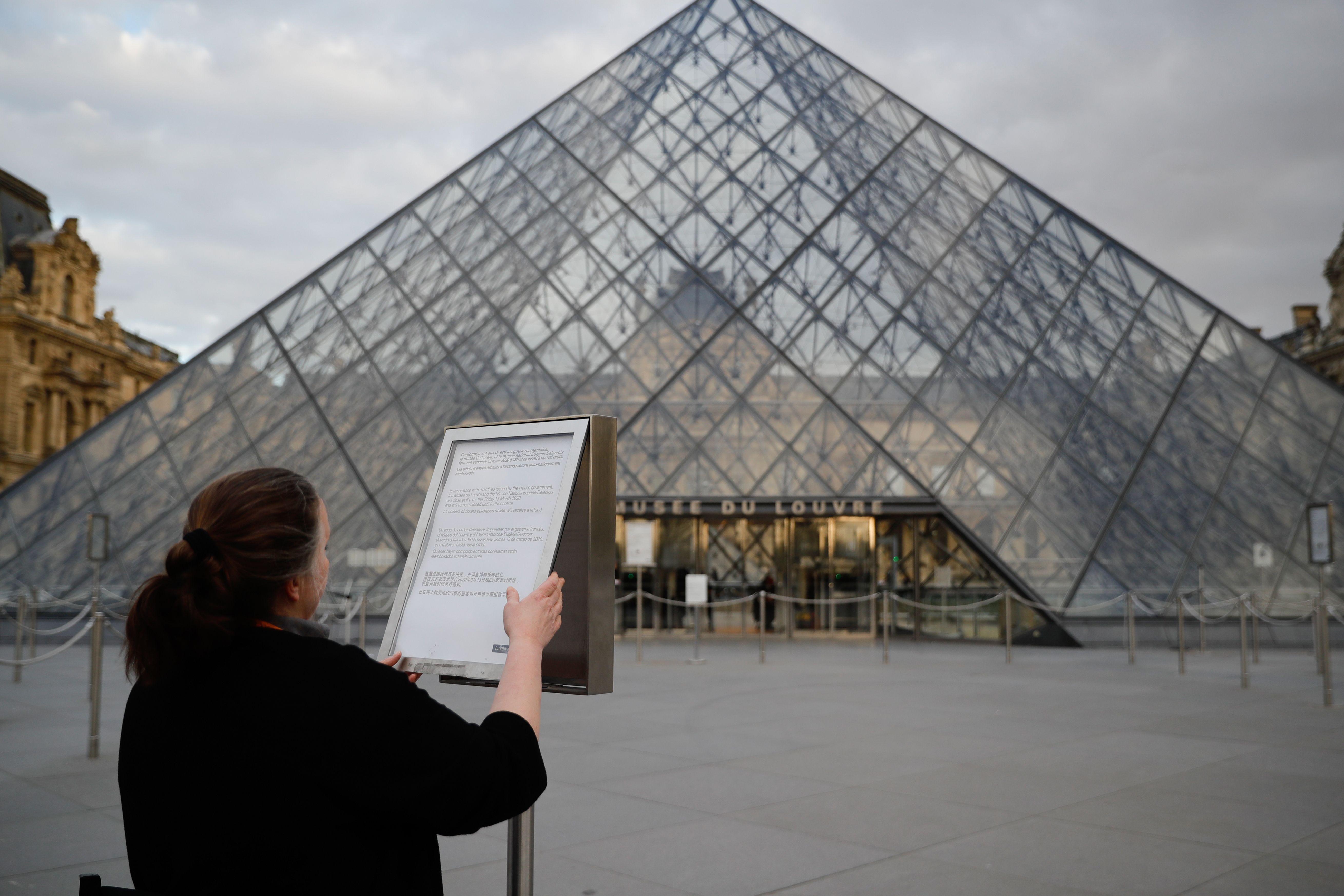 A tourist reads an information sign in front of the Louvre pyramid designed by Ieoh Ming Pei at the entrance of the Musee du Louvre in Paris, undefinitely closed to the public amid concerns on the COVID-19 outbreak, caused by the novel coronavirus, on March 13, 2020. - The Louvre in Paris, the world's most visited museum, said on March 13, 2020, that it was closing "until further notice" because of the coronavirus. The closure of the museum, which had 9.6 million visitors last year, came after the French government banned all gatherings of over 100 people to limit the spread of the virus. The Palace of Versailles -- France's other big tourist attraction with nearly 10 million tourists a year -- swiftly followed suit. (Photo by Thomas SAMSON / AFP) / RESTRICTED TO EDITORIAL USE - MANDATORY MENTION OF THE ARTIST UPON PUBLICATION - TO ILLUSTRATE THE EVENT AS SPECIFIED IN THE CAPTION