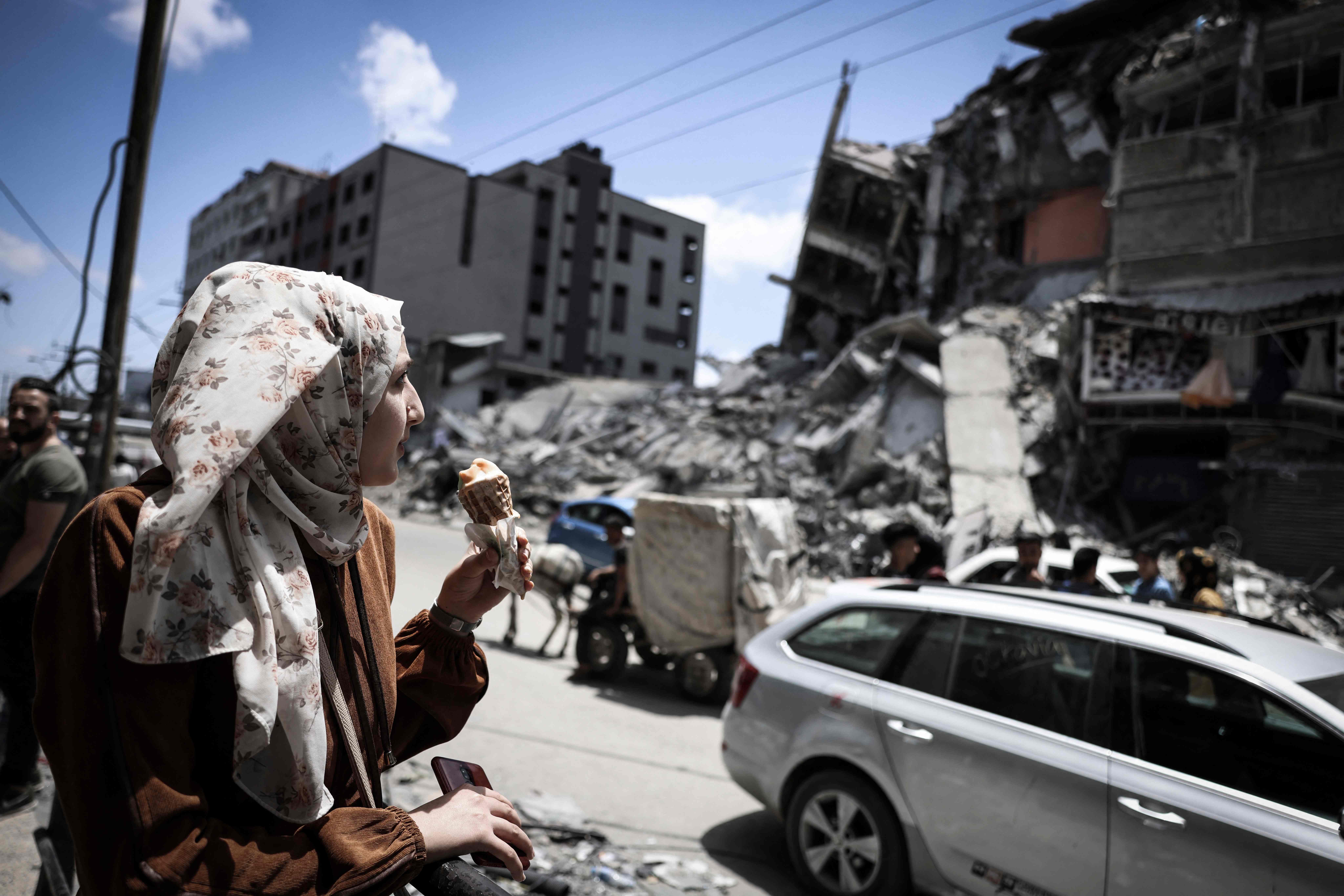 TOPSHOT - A Palestinian woman eats ice-cream in front of the destroyed Al-Shuruq building, destroyed by an Israeli air strike, on May 22, 2021, in Gaza City. - A ceasefire between Israel and Hamas, the Islamist movement which controls the Gaza Strip, appeared to hold today after 11 days of deadly fighting that pounded the Palestinian enclave and forced countless Israelis to seek shelter from rockets. (Photo by MAHMUD HAMS / AFP)