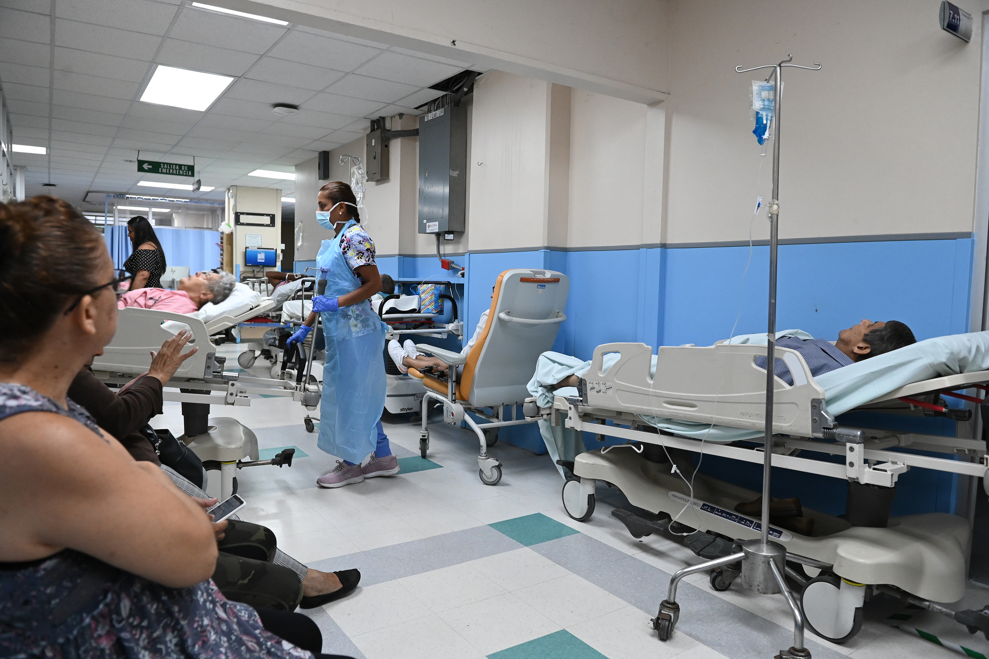 Salón de emergencias de un hospital con pacientes esperando en sillas de ruedas y camillas.