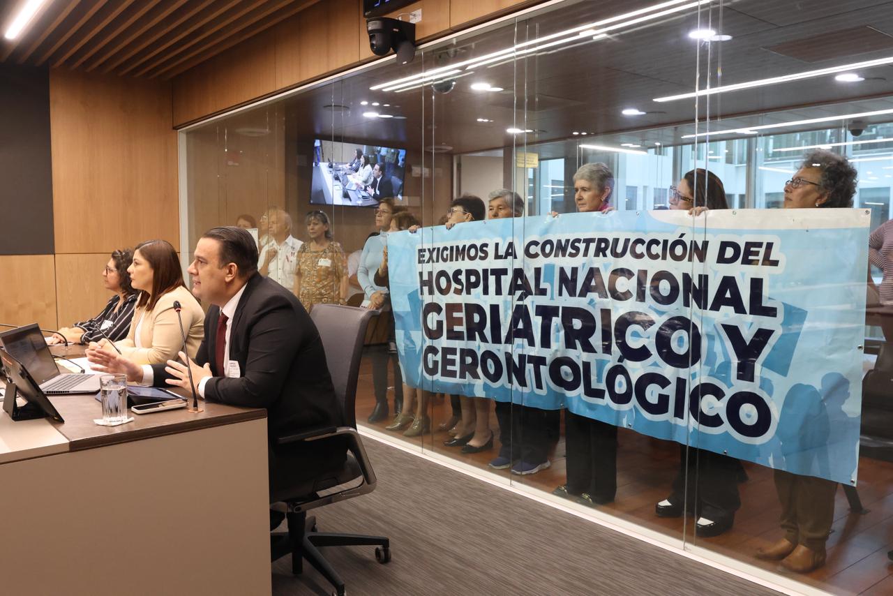 Se ve una sala de audencias, hay tres comparecientes y detrás de un vidrio un grupo del público sostiene una manta donde pide la construcción del hospital nacional de geriatría.