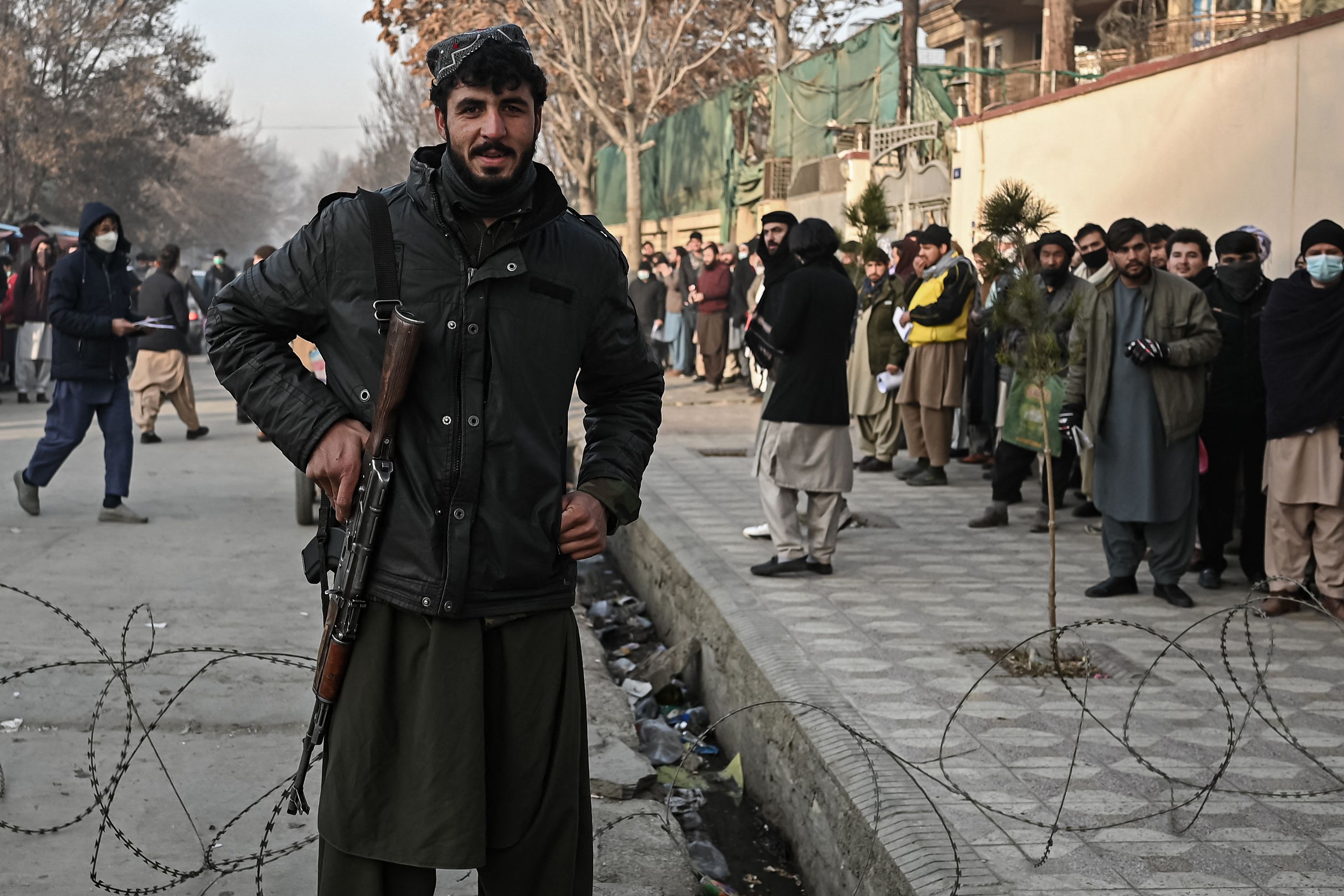 A Taliban fighter stands guard at a checkpoint as people queue to enter the passport office in Kabul on December 19, 2021, after Afghanistan's Taliban authorities said they will resume issuing passports. (Photo by Mohd RASFAN / AFP)