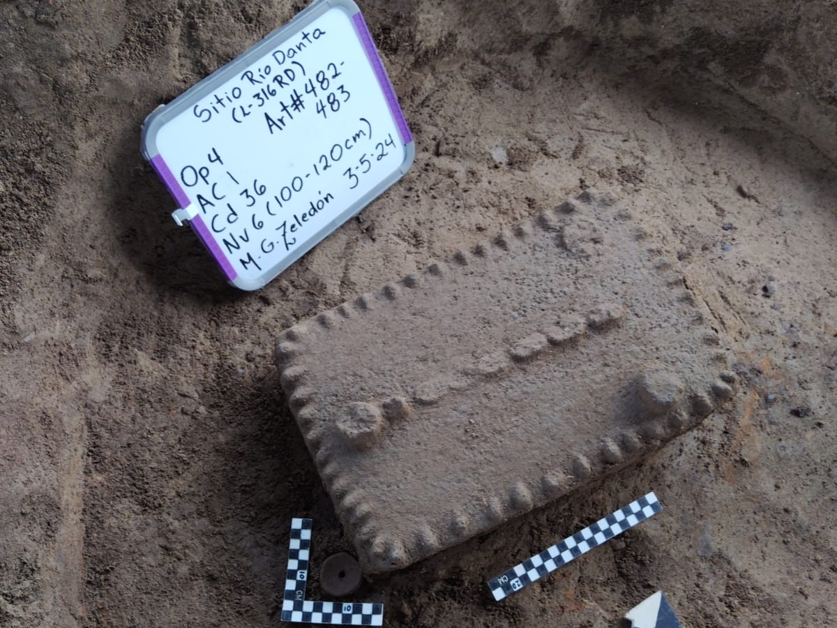 Un metate en un terreno excavado. Atrás se ven las escalas y guías de arqueología.