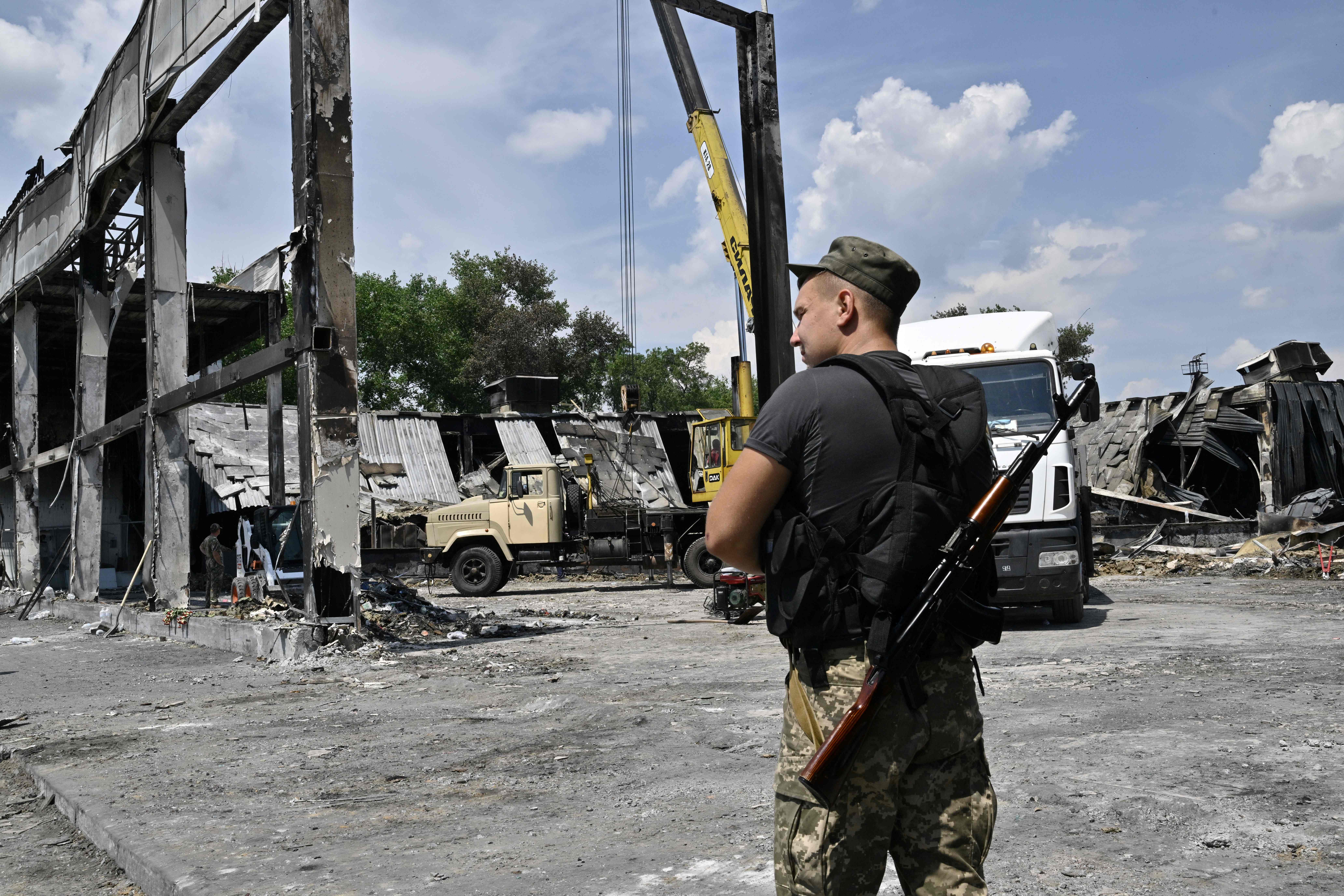 A Ukrainian serviceman stands alert as workers clear rubble and debris from The Amstor Mall in Kremenchuk, on June 29, 2022, two days after it was hit by a Russian missile strike according to Ukrainian authorities. - A missile strike on a crowded mall in Kremenchuk, central Ukraine killed at least 18 people. (Photo by Genya SAVILOV / AFP)
