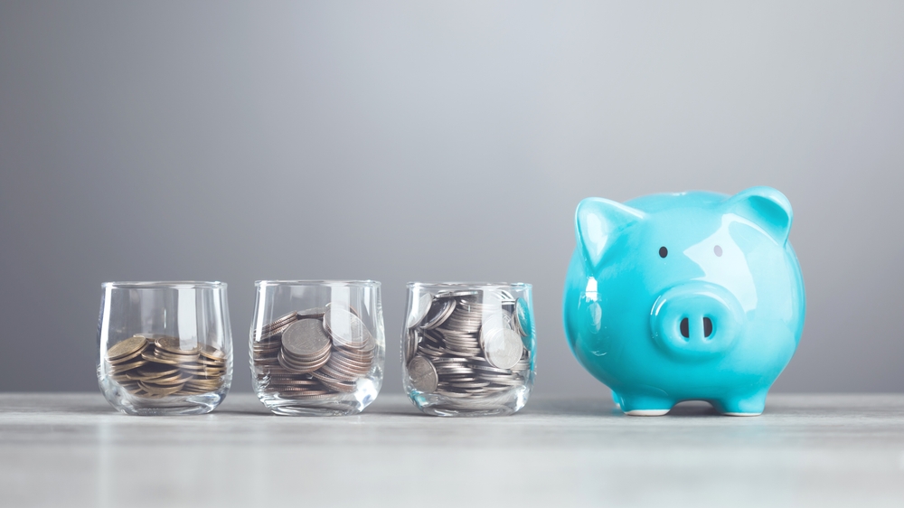 Alcancía azul junto a tres vasos con monedas que representan opciones de inversión y diversificación de capital.