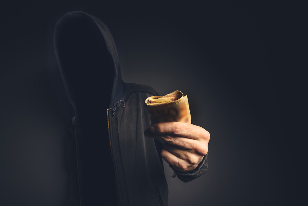 Unrecognizable hooded computer hacker offering cash money, cyber crime, blackmail and extortion concept.
