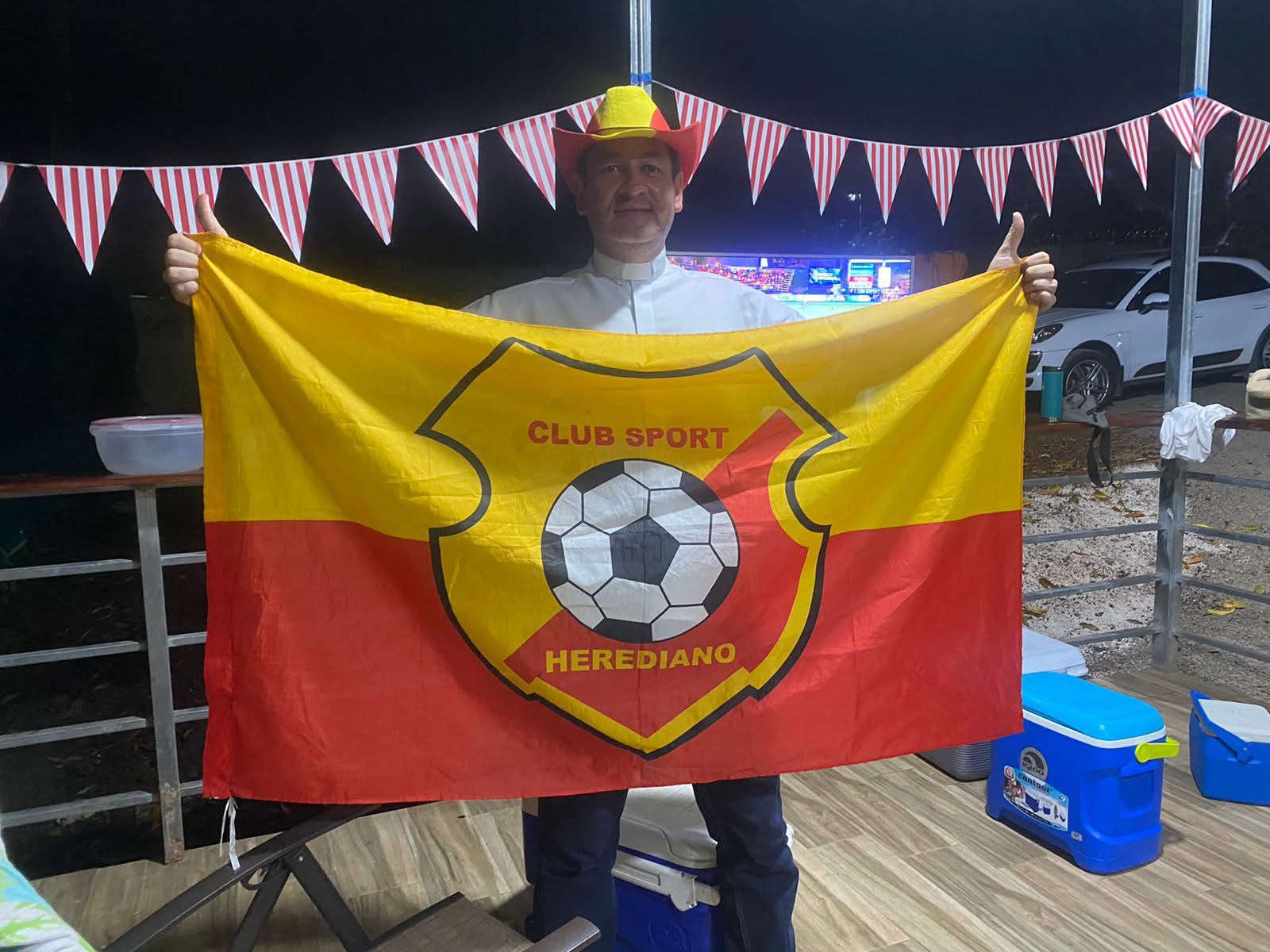 Sacerdote sostiene por las puntas una gran bandera mitad amarilla y mitad roja con el escudo de Herediano en el centro. Además usa un sombrero estilo vaquero, de color rojo en los costados y amarillo en el centro.