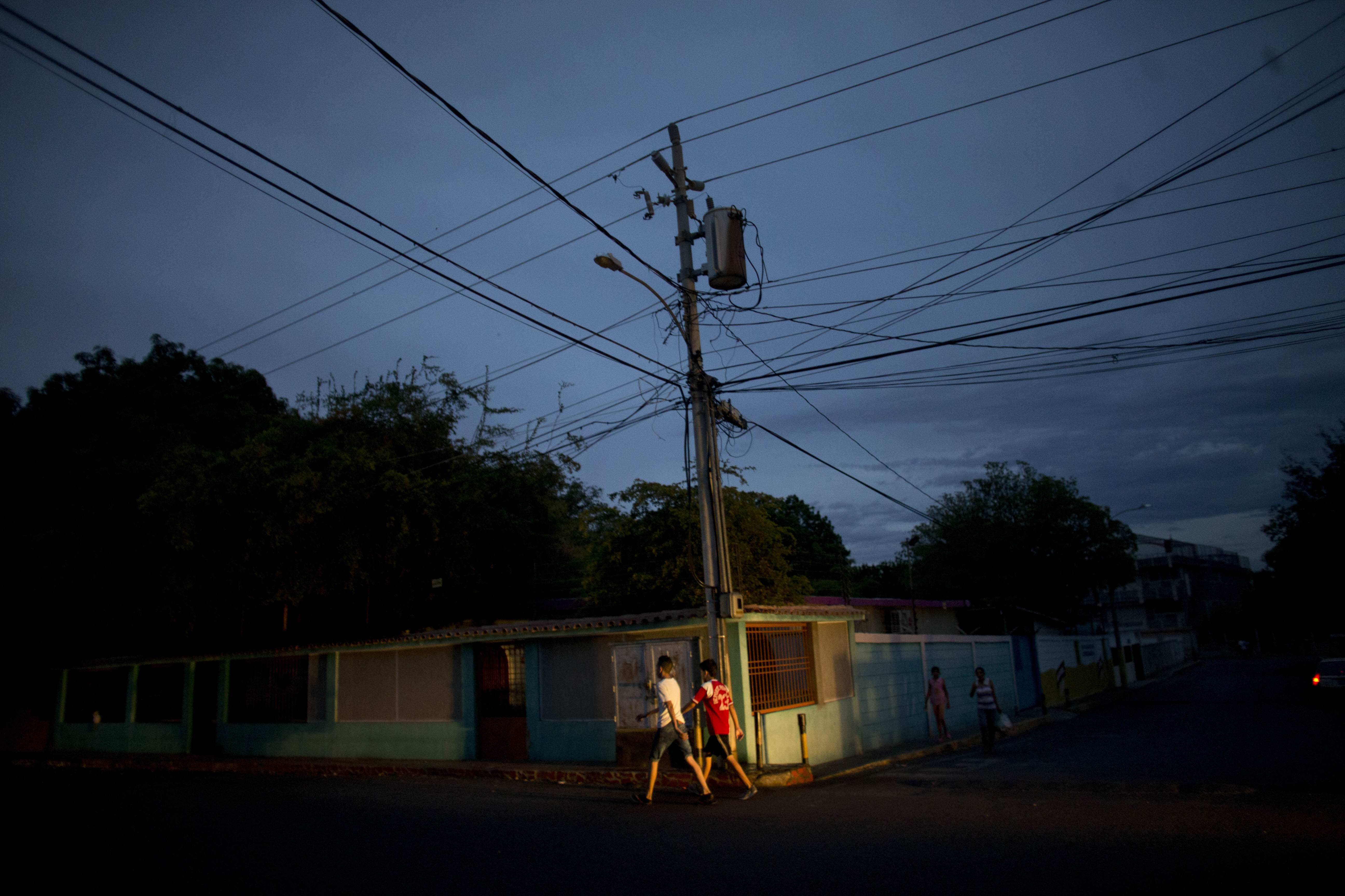 In this April 18, 2018 photo, residents walk under a charred transformer box hanging on a pole that exploded in late March, in La Chinita neighborhood in Maracaibo, Venezuela. Officials repeatedly promised the parts needed to repair it would arrive the next day. So far they haven't come. (AP Photo/Ariana Cubillos)