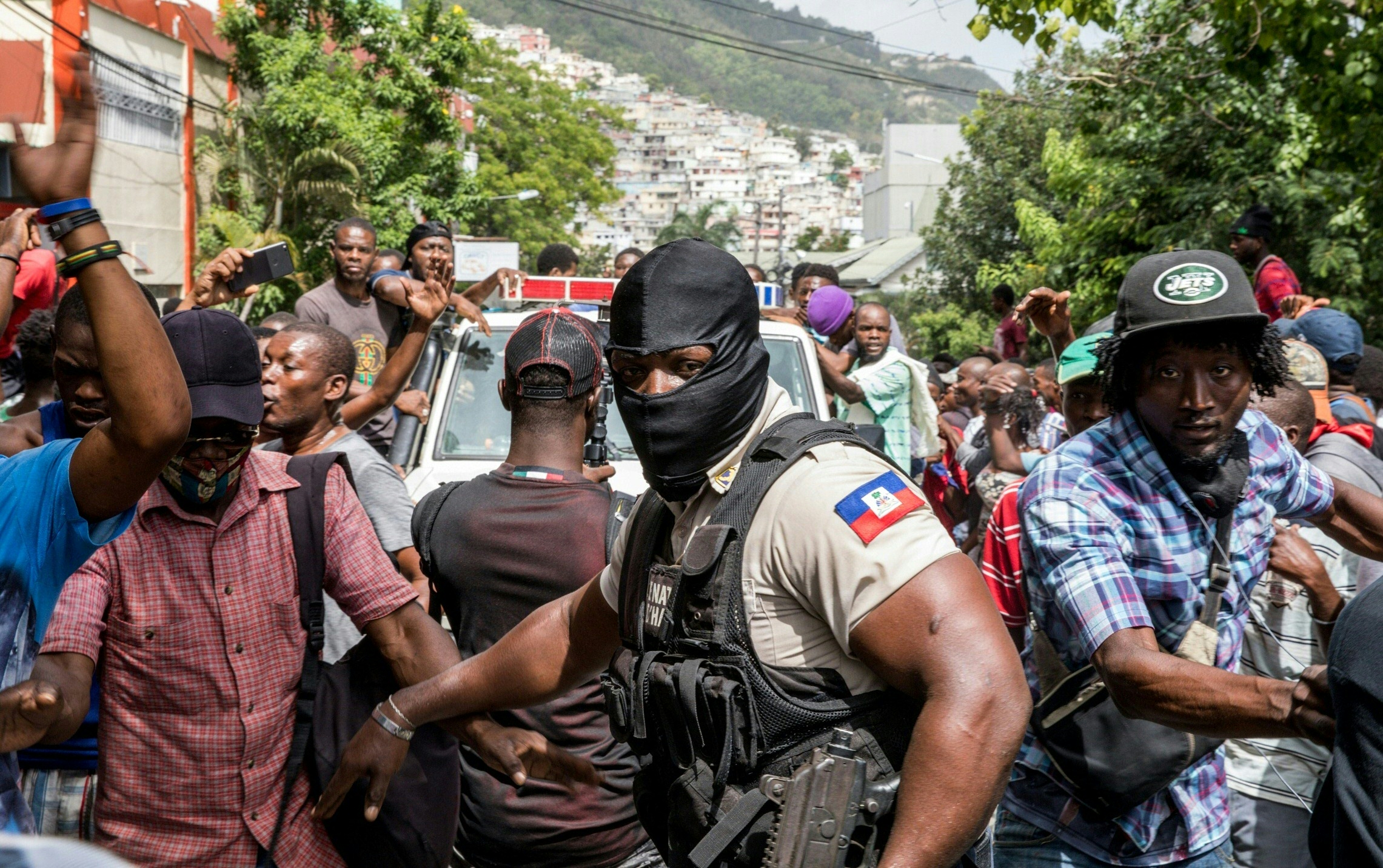 People cheer as a police car drives past at the Petionville Police station where armed men, accused of being involved in the assassination of President Jovenel Moise, are being held in Port au Prince on July 8, 2021. - Police in Haiti have surrounded a group of possible suspects in the assassination of President Jovenel Moise, the UN envoy to Haiti said. Helen La Lime said from the Haitian capital that four members of a group that attacked the presidential palace Wednesday and shot the president have been killed by police and six others are in custody. (Photo by Valerie Baeriswyl / AFP)