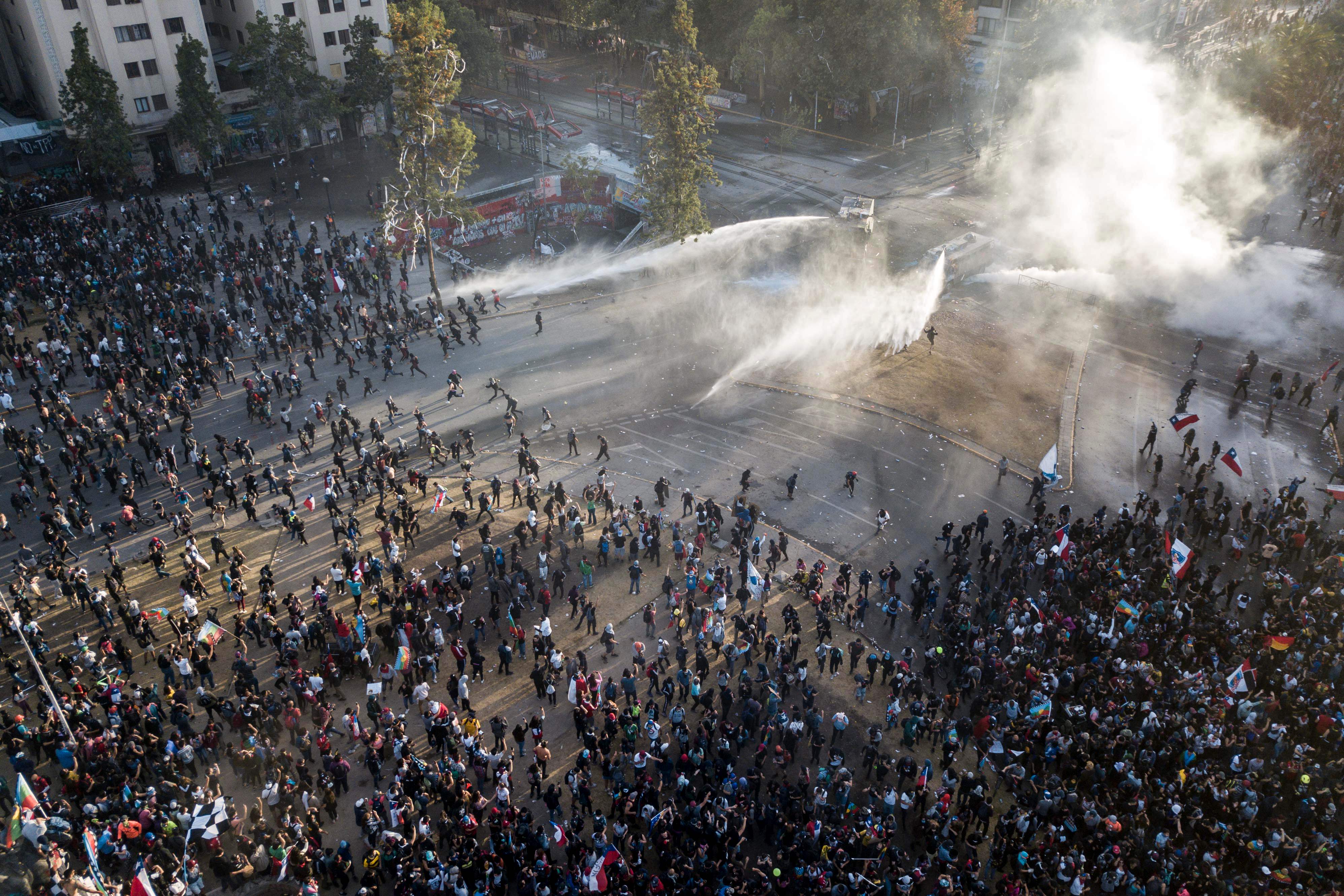 Aerial view of security forces spraying water as people take part in the so-called 'Mourning March' in Santiago on November 01, 2019, to protest against the death of 23 people after more than ten days of civil unrest. - Chile's government met with opposition leaders Thursday in a fresh bid to end deadly protests that forced the country to abandon hosting two major economic and climate summits, but leftist parties poured scorn on the efforts. The unrest started with protests against a rise in transport tickets and other austerity measures and descended into vandalism, looting, and clashes between demonstrators and police. (Photo by JAVIER TORRES / AFP)