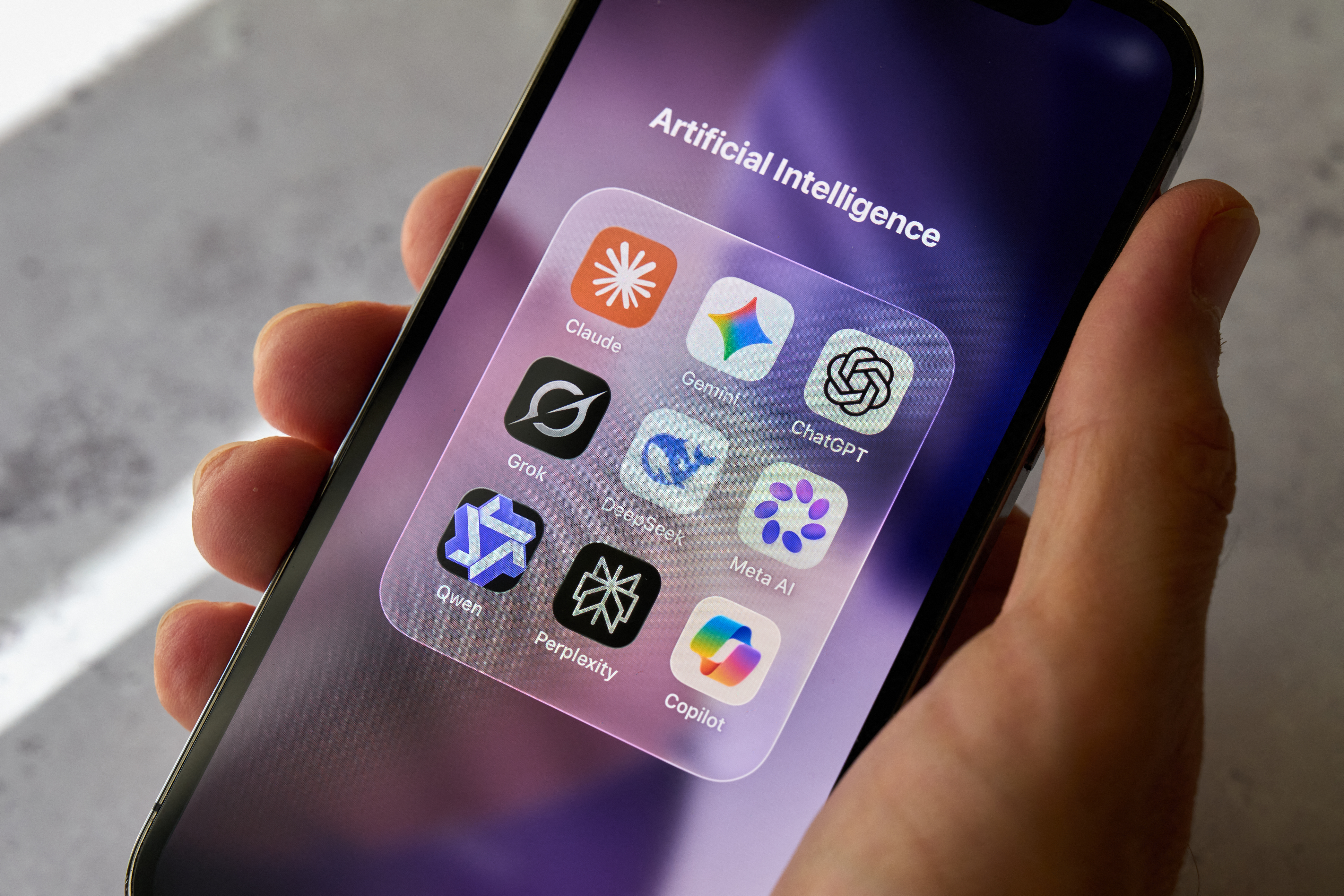 Una mano sostiene un teléfono inteligente que muestra una carpeta de aplicaciones de inteligencia artificial, entre ellas Claude, Gemini, ChatGPT, Grok, DeepSeek, Meta AI, Qwen, Perplexity y Copilot. La fotografía fue tomada en Italia el 5 de julio de 2026.