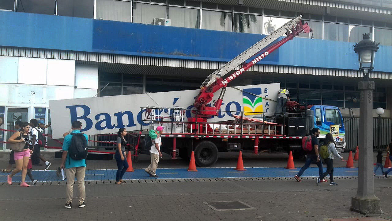 Operarios desmontan el rótulo de Bancrédito en un edificio en Costa Rica, mientras transeúntes caminan por la acera. La imagen refleja el cierre de la entidad en el marco de la reducción del número de bancos en el país.