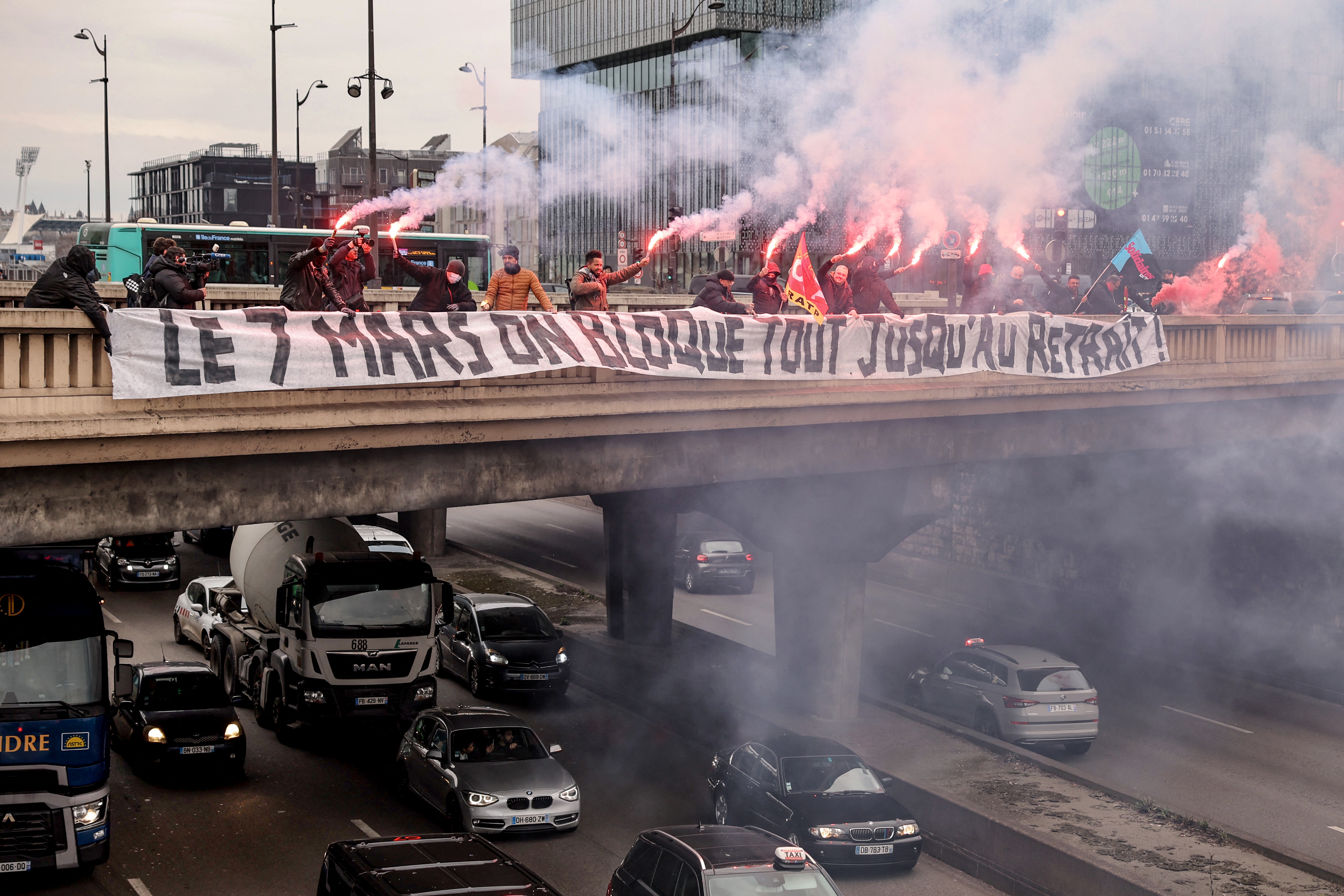 Los conductores de autobuses de la estatal RATP, que opera el gran sistema de transporte de París, encienden bengalas detrás de una pancarta que dice en francés "el 7 de marzo, bloqueemos todo hasta la retirada" en un puente sobre la carretera de circunvalación de París.
