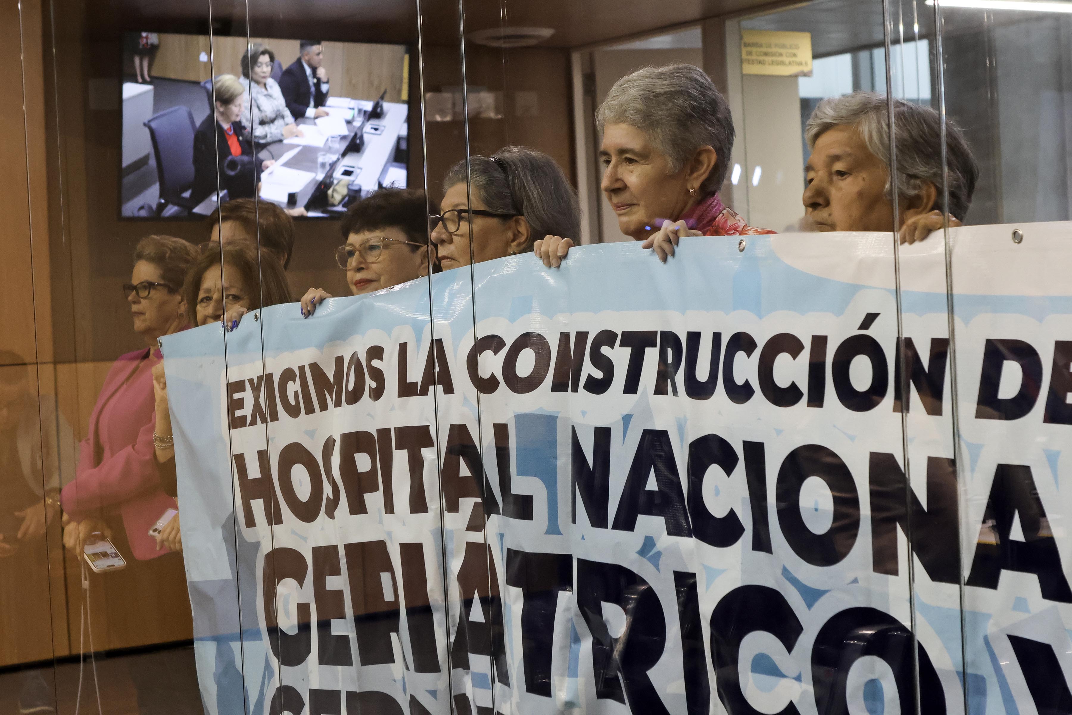 Personas adultas mayores sostienen una manta que pide construir un nuevo hospital.