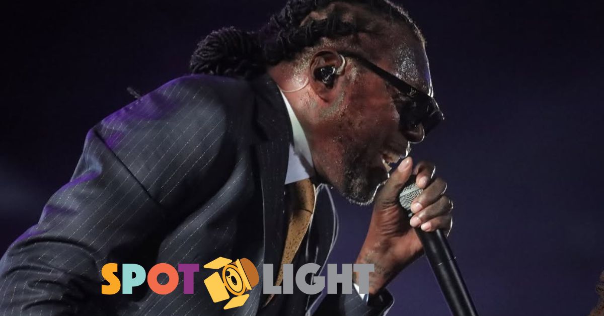 Vybz Kartel cantando en concierto, con el micrófono en mano. usa un traje de color negro y corbata dorada.