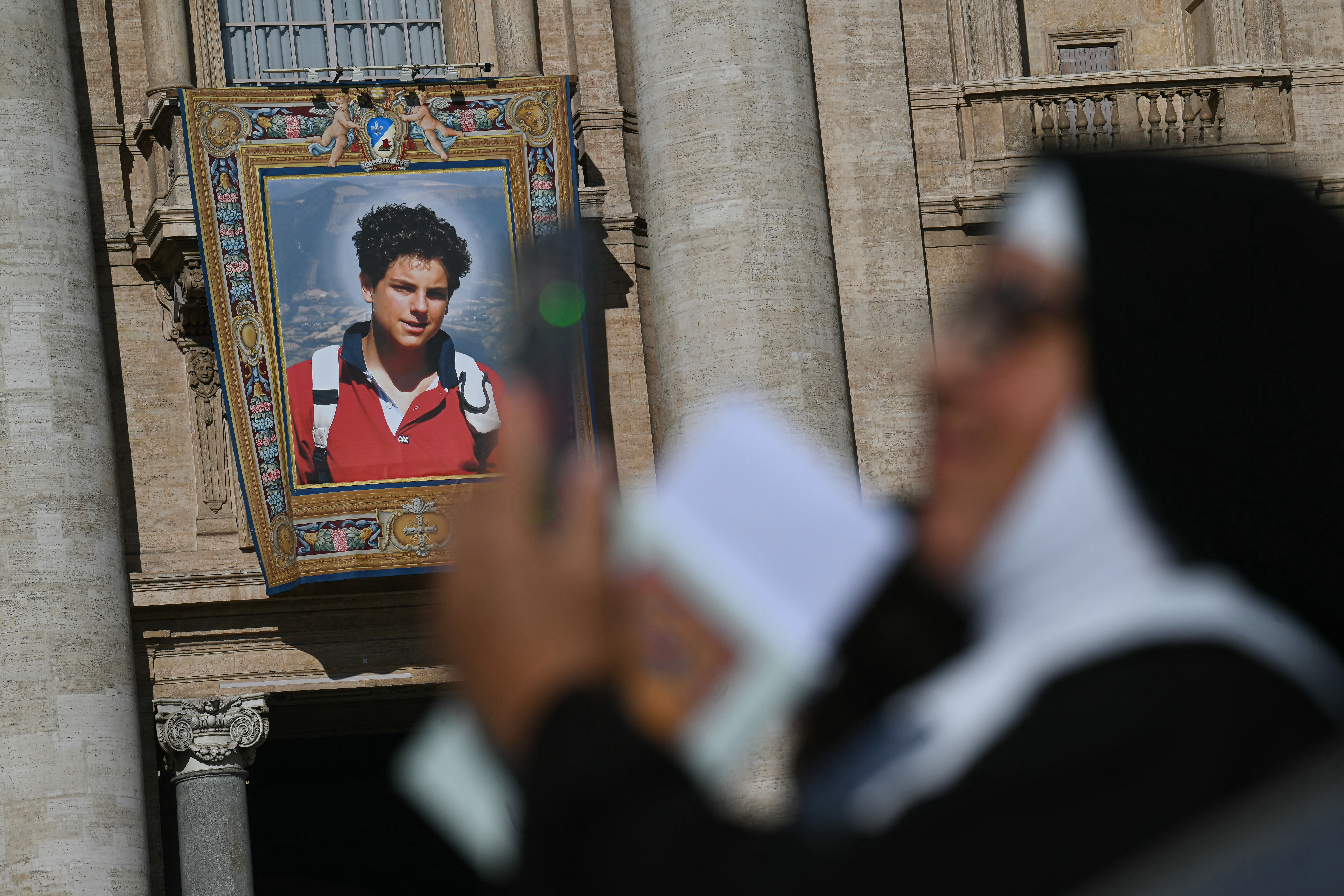 A nun checks her phone as a portrait of late Italian teenager Carlo Acutis is displayed onto the facade of Saint Peter's Basilica in the background ahead of a Holy Mass and canonisation of Blessed Carlo Acutis and Pier Giorgio Frassati in St Peter's Square at the Vatican on September 7, 2025. An Italian teenager dubbed "God's Influencer" for his efforts to spread the Catholic faith online will become the first millennial saint Sunday at a canonisation attended by thousands of pilgrims. Computer whiz Carlo Acutis, who died of leukemia in 2006 aged 15, will be raised to sainthood by Pope Leo XIV in a solemn ceremony in St Peter's Square at the Vatican. Italian Pier Giorgio Frassati, a mountaineering enthusiast who died in 1925 and was known for his social and spiritual commitment, will also be made a saint on September 7, 2025. (Photo by Filippo MONTEFORTE / AFP)