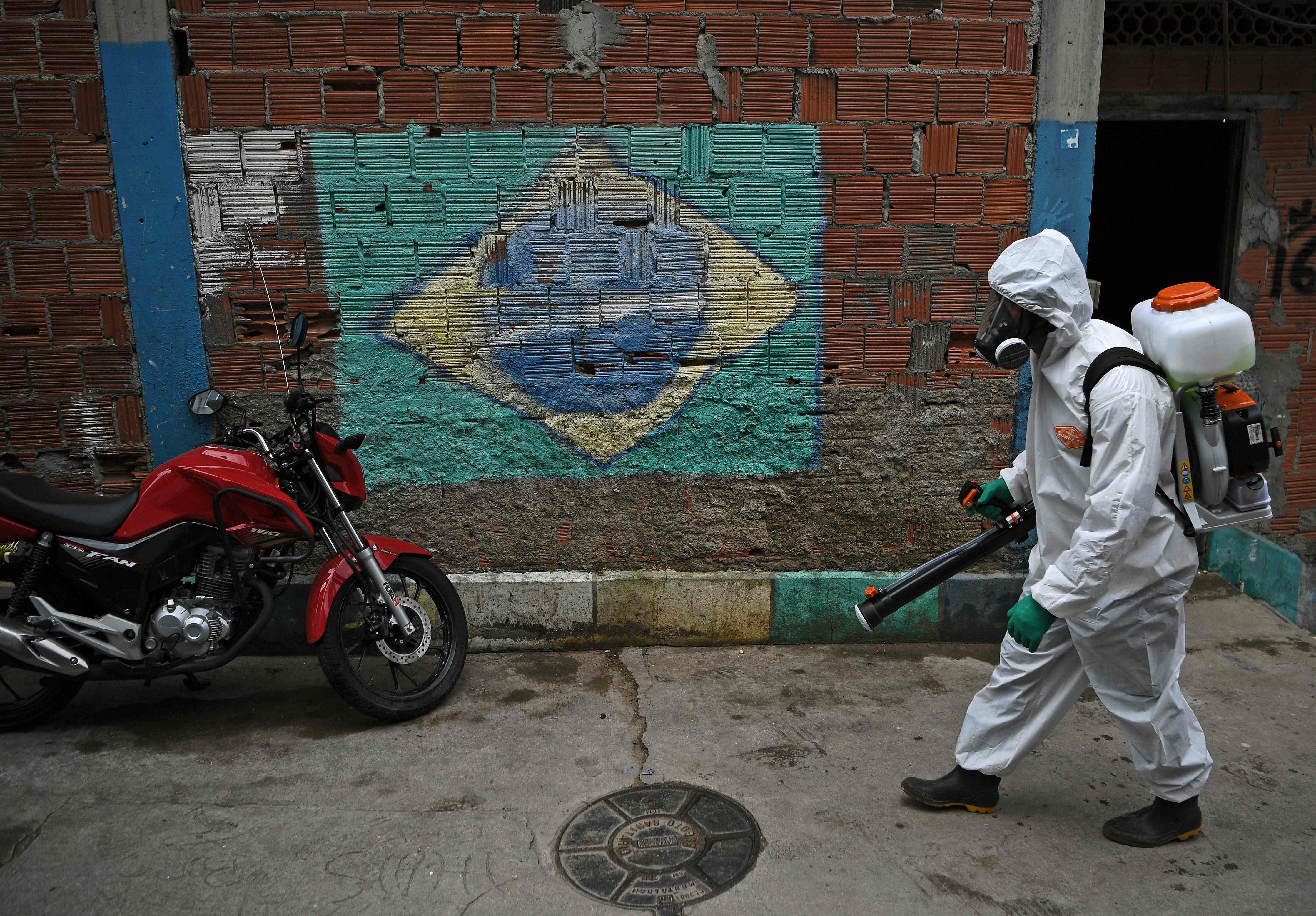 A volunteer walks past a mural of the Brazilian flag as he disinfects an area inside Babilonia favela, in Rio de Janeiro on April 18, 2020. - Neighbours of the Santa Marta favela are helping to disinfect and train volunteers in Babilonia favela to fight against COVID-19 coronavirus. Favela residents are taking matters into their own hands and providing services during the pandemic because they claim that they are not being assisted by the government. (Photo by CARL DE SOUZA / AFP)