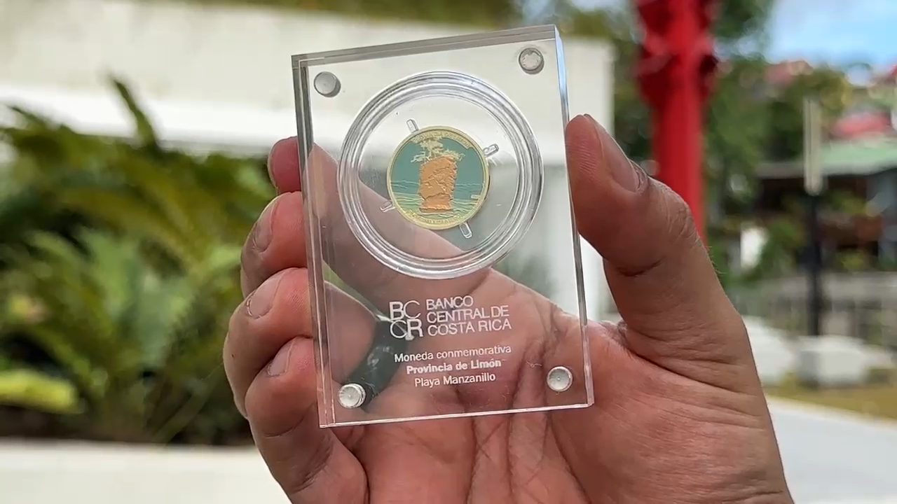 La mano de un hombre con traje sostiene una moneda coleccionable de 25 colones con la imagen de Playa Manzanillo.