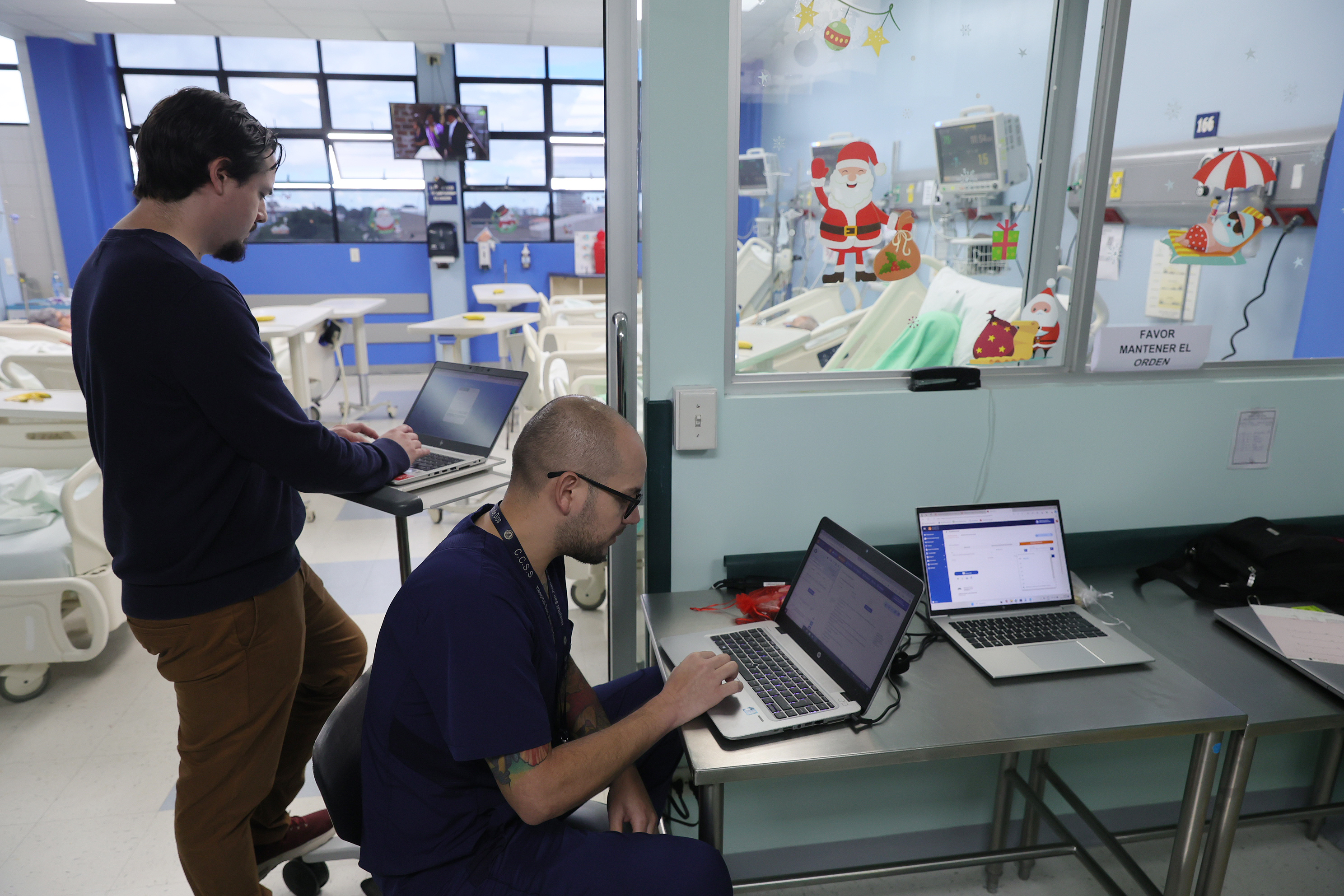 Salón de hospital con camas de hospitalización al fondo y dos médicos en primer plano con computadoras usando expediente digital en salud de CCSS.