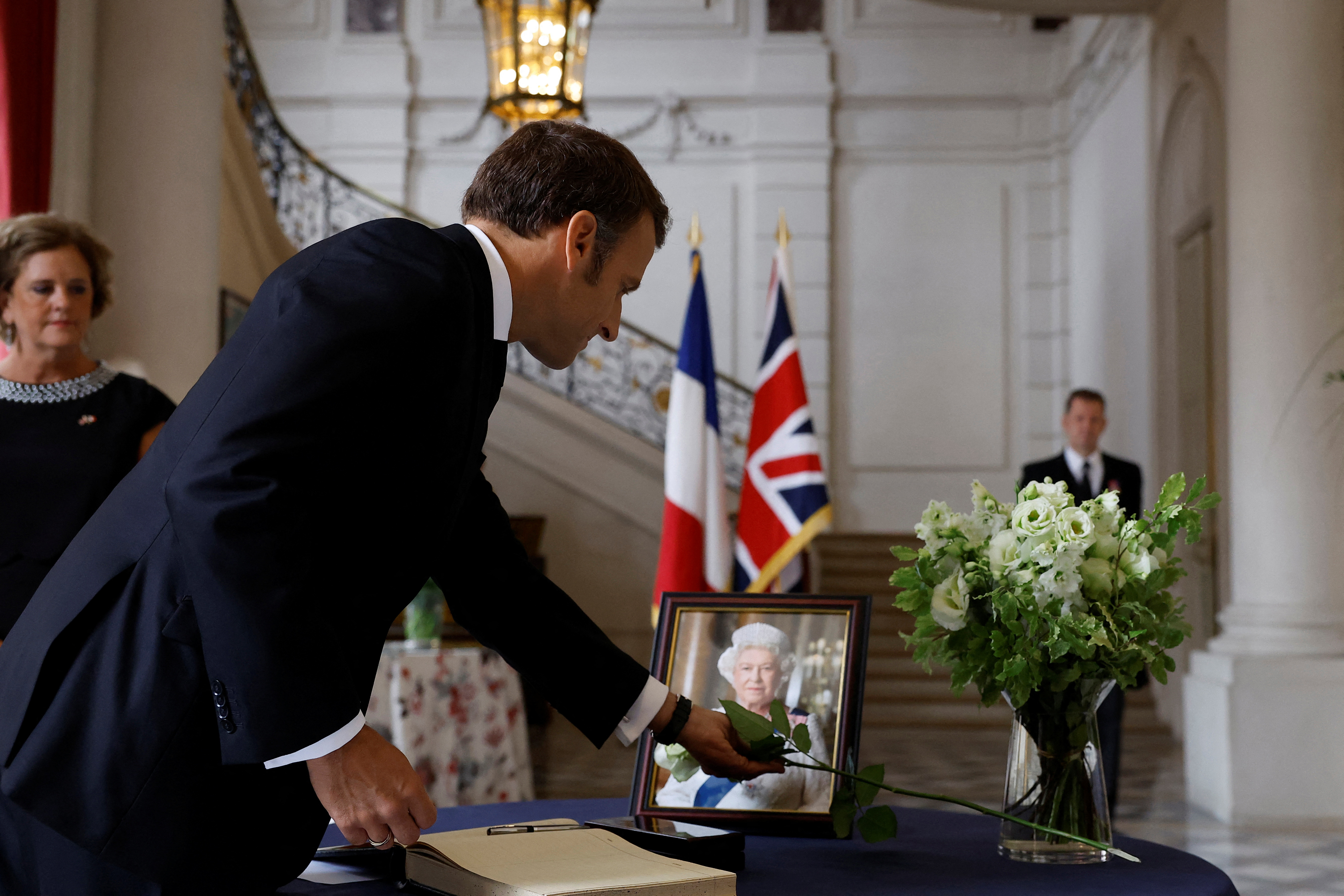 French President Emmanuel Macron places a white rose next to a portrait of Queen Elizabeth as British Ambassador to France Menna Rawlings looks on during the signature of a condolence book, following the passing of Britain's Queen Elizabeth, at the British Embassy in Paris, France, September 9, 2022. REUTERS/Christian Hartmann/Pool