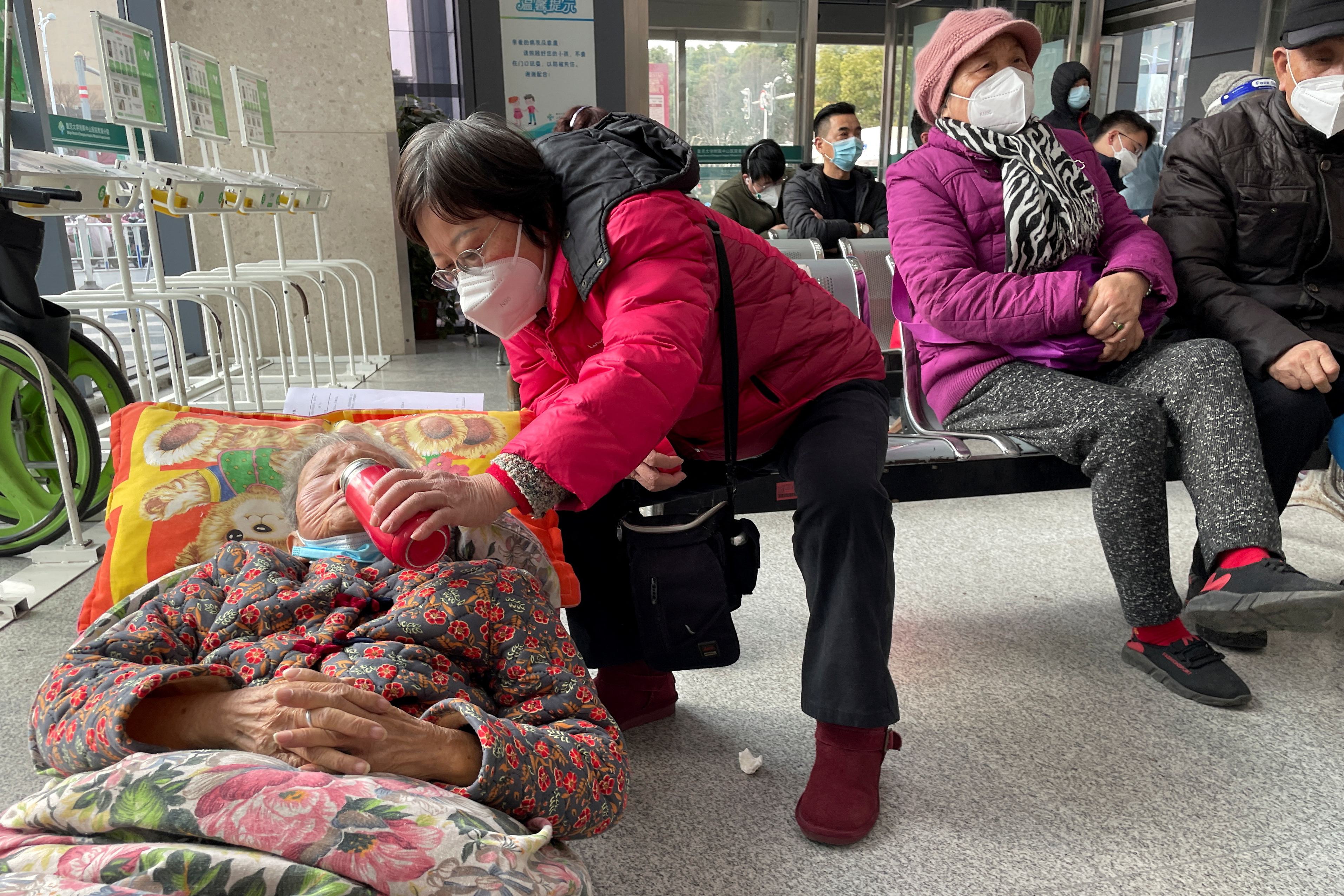 Una mujer le da de beber a un anciano acostado en una camilla mientras espera en el departamento de emergencias de un hospital, en medio del brote de la enfermedad por coronavirus (COVID-19) en China