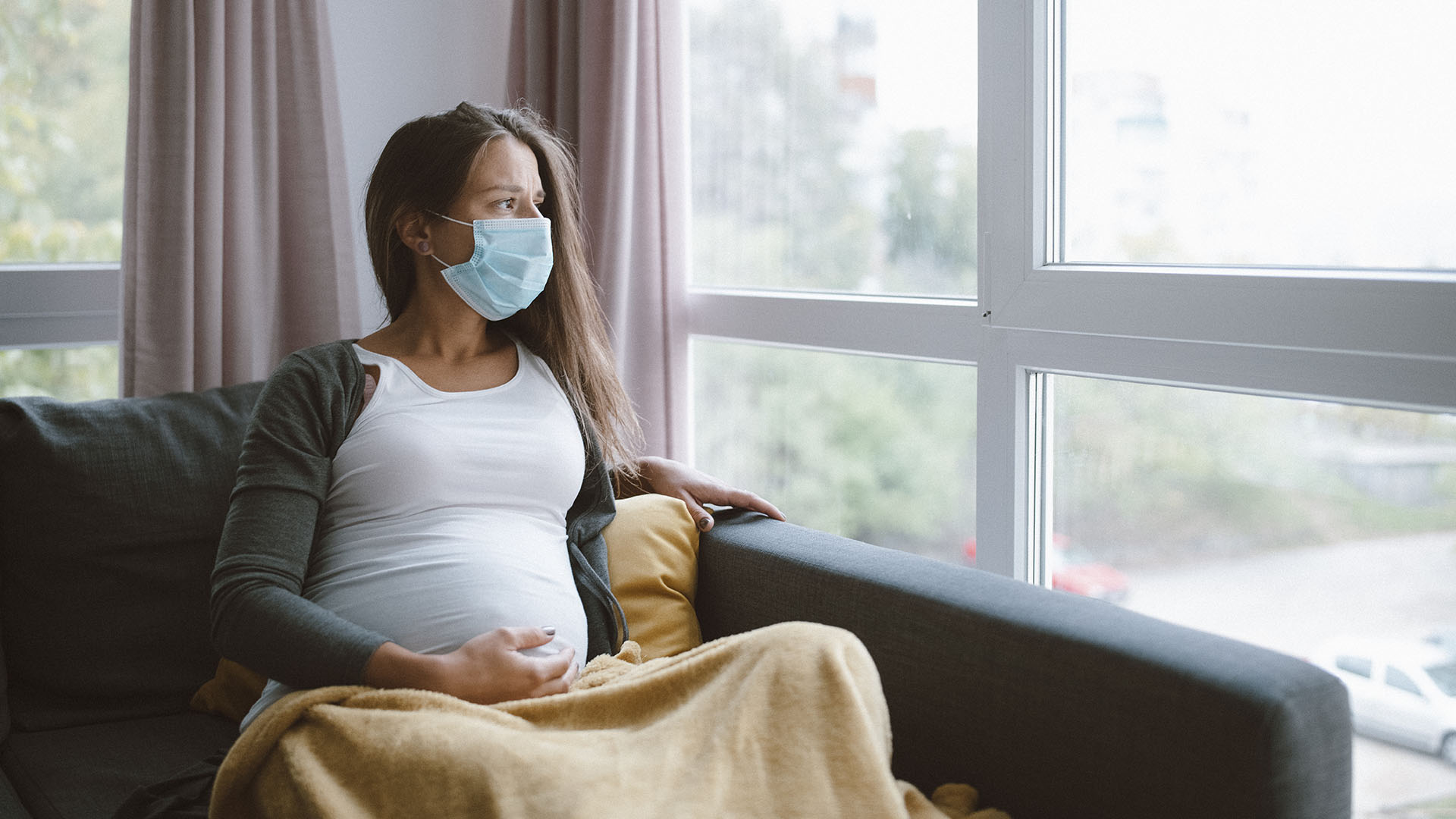 worried pregnant woman sitting at home with protective face mask and looking through the window. (Getty)